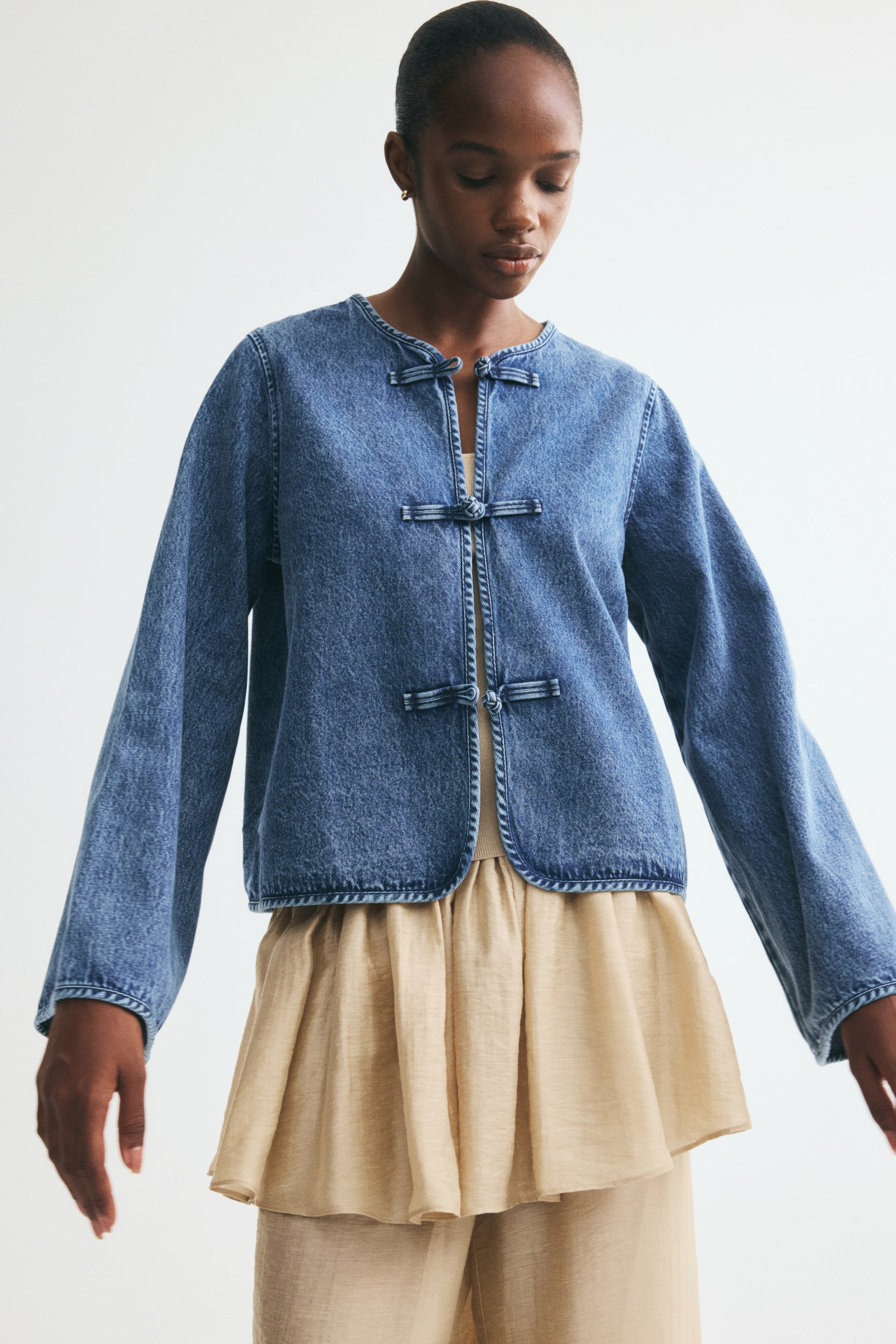 View larger image: The model is wearing a short cotton denim jacket. It features a round neckline and three pairs of knot buttons down the front, with the jacket styled open to reveal a light-coloured garment underneath.