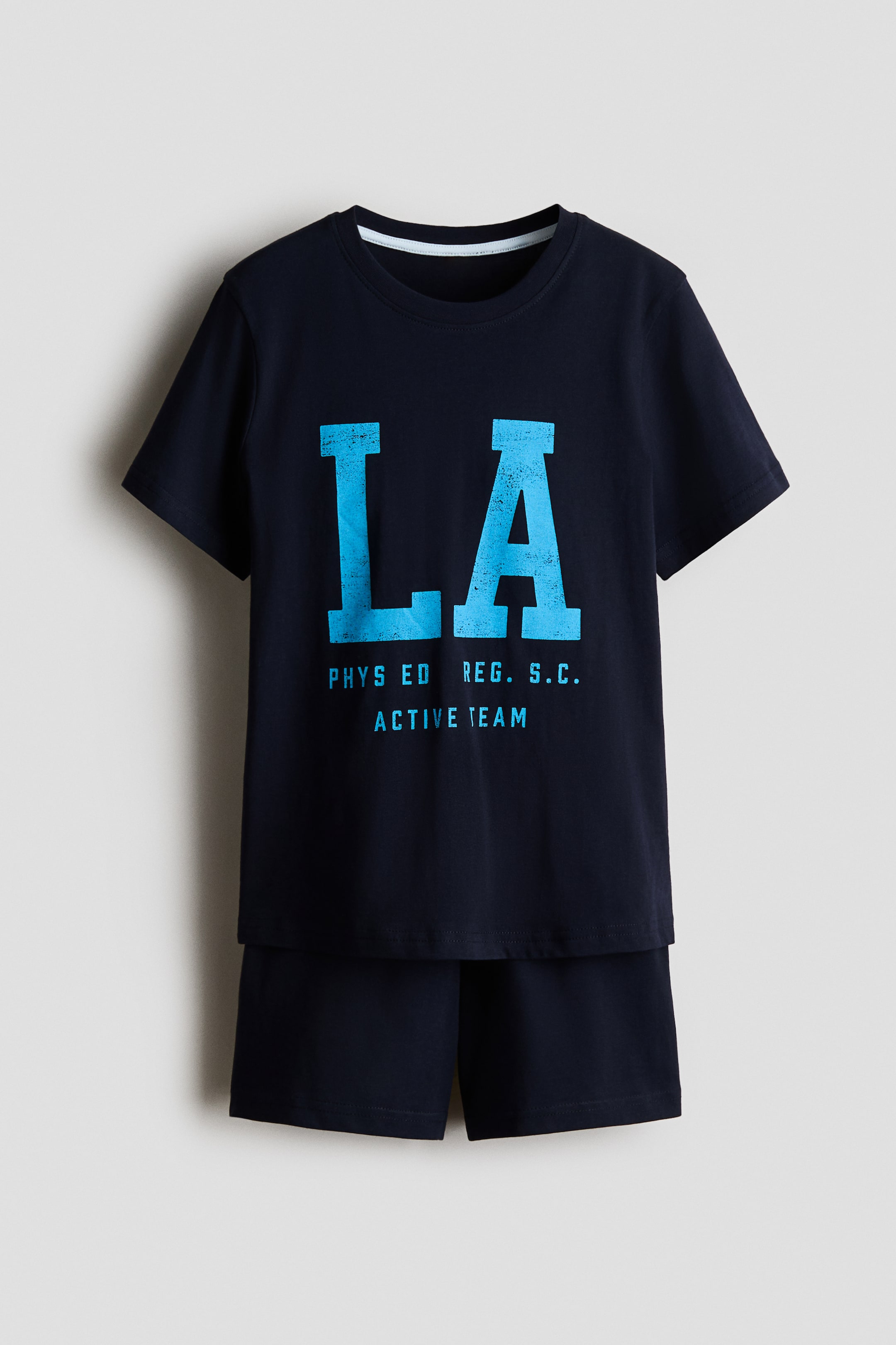 View larger image: Set baju tidur kanak-kanak berwarna biru gelap ini menampilkan t-shirt berkolar bulat dengan lengan pendek dan seluar pendek yang sepadan, dipersembahkan menghadap ke depan. T-shirt ini mempamerkan grafik "LA" besar berwarna biru muda yang kelihatan usang dengan teks lebih kecil "PHYS ED REG. S.C. ACTIVE TEAM" di bahagian hadapan.