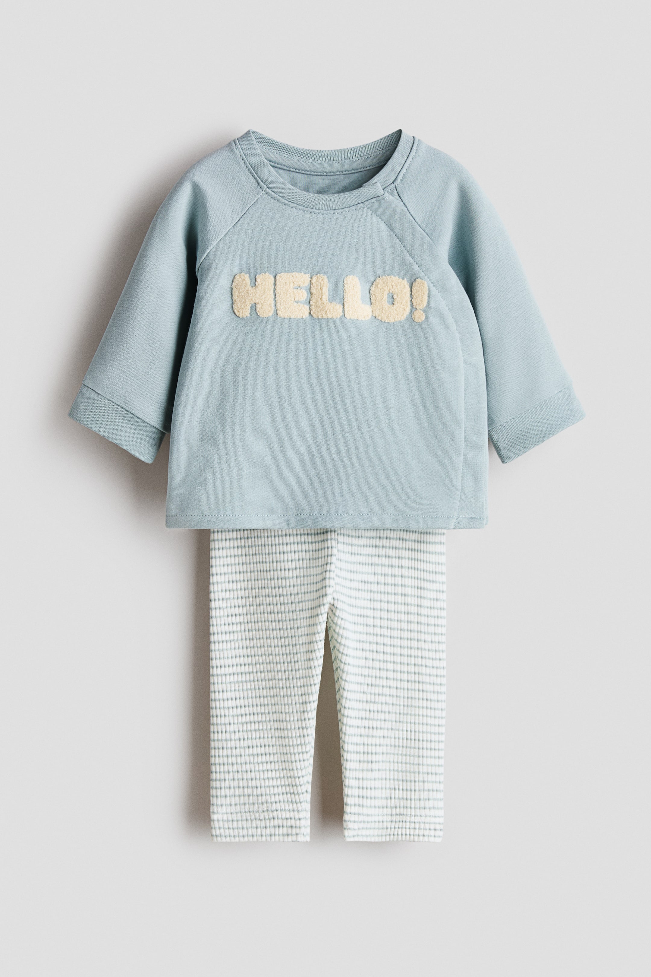 Vis større bilde: Et sett bestående av en lyseblå topp og ribbestrikkede leggings vises forfra. Toppen har lange raglanermer og en lodden, lystfarget 'HELLO!'-påskrift, mens leggingsene har et rutenettmønster i hvitt og lyseblått.