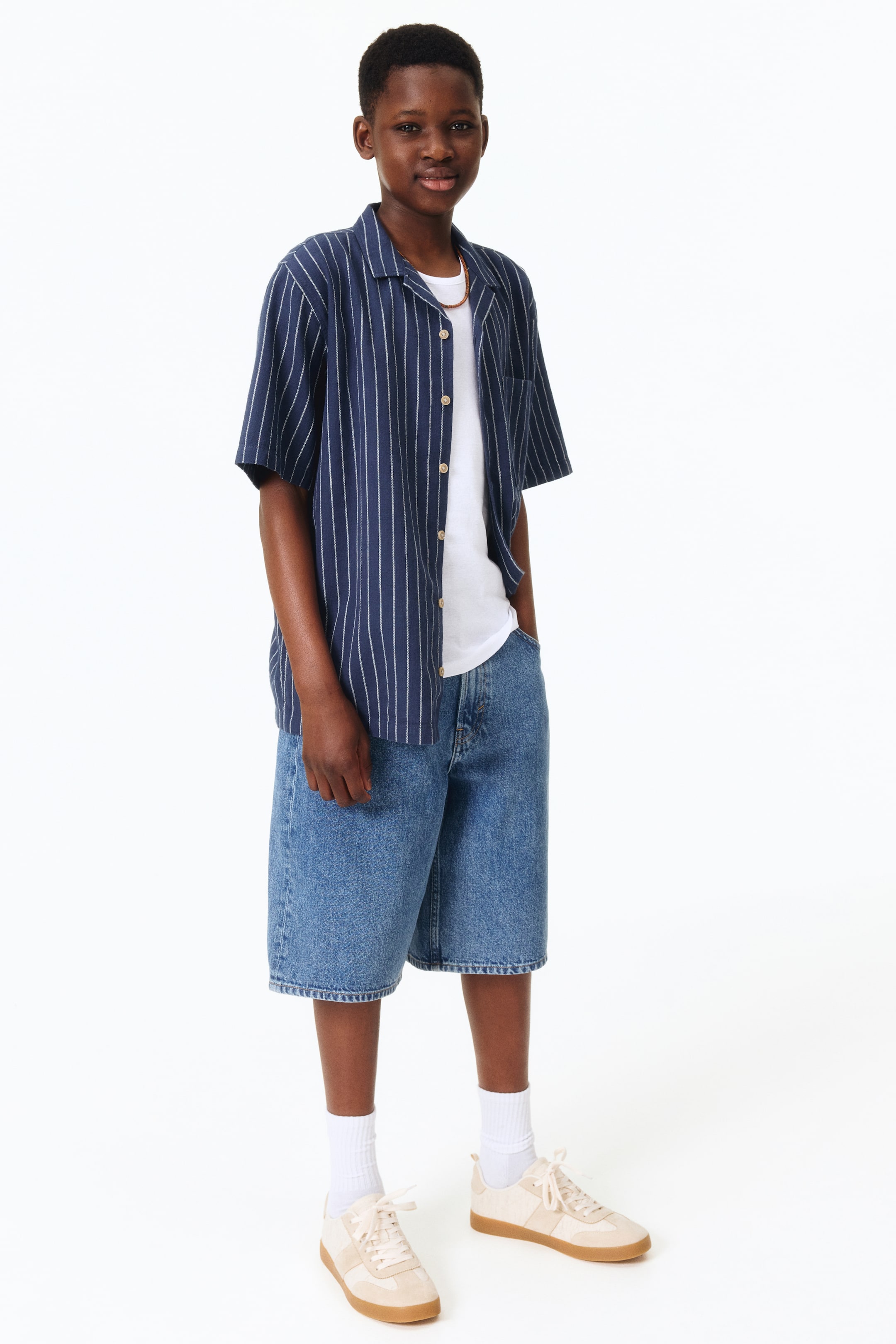 Voir une image plus grande: Une chemise bleu marine à manches courtes, ornée de rayures verticales blanches et d'un col cubain, est portée déboutonnée sur un t-shirt blanc à col rond. Un short en denim bleu à hauteur des genoux complète le look, accompagné de chaussettes blanches côtelées et de baskets basses beige et crème à lacets avec semelles en gomme, le tout présenté de face.