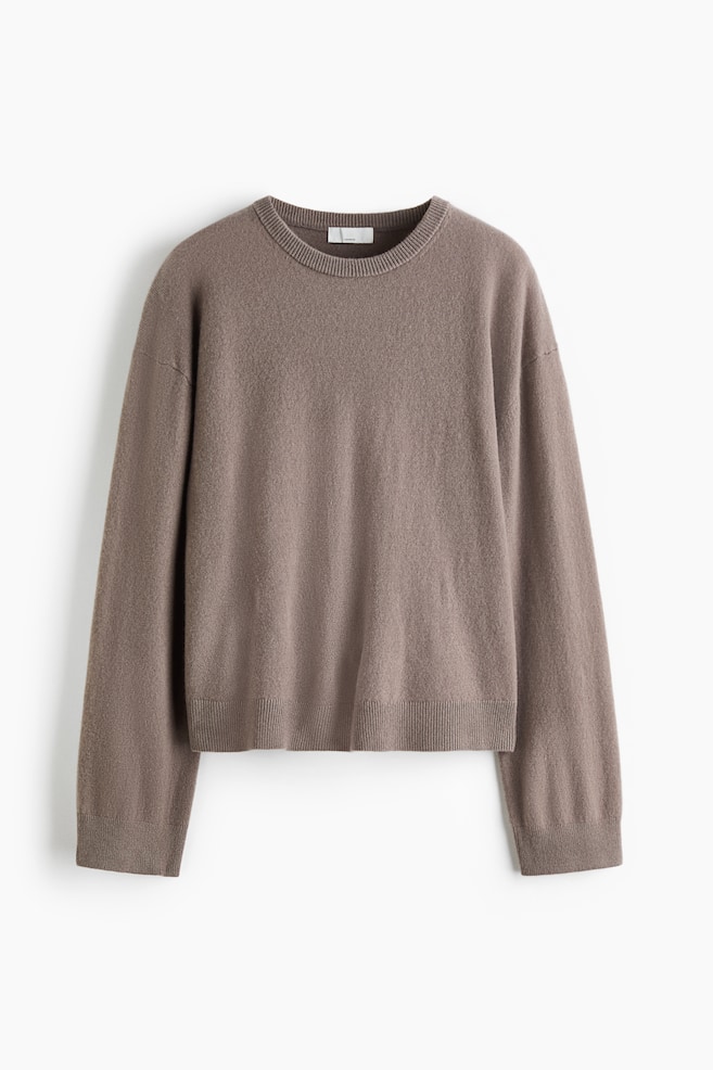 A relaxed-fit, taupe-brown cashmere jumper is shown from the front, featuring a round, rib-trimmed neckline and low dropped shoulders. The long-sleeved garment displays a soft, fine-knit texture with ribbed detailing at the cuffs and hem. - 1