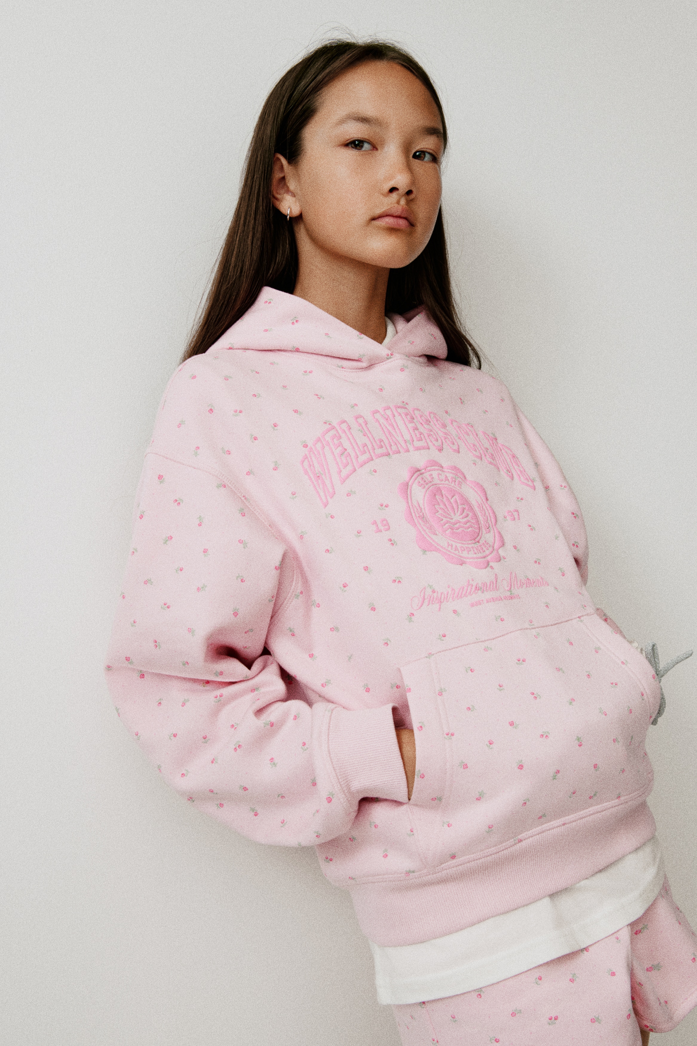 En frontvendt lys rosa oversized hettegenser har et heldekkende mini blomsterprint og en sentral grafikk med teksten Â«WELLNESS CLUBÂ» over et sirkelformet emblem. Hettegenseren har en omslagshette foran, droppede skuldre, kengurulomme og brede ribbestrikkede avslutninger ved mansjettene og nederkanten.