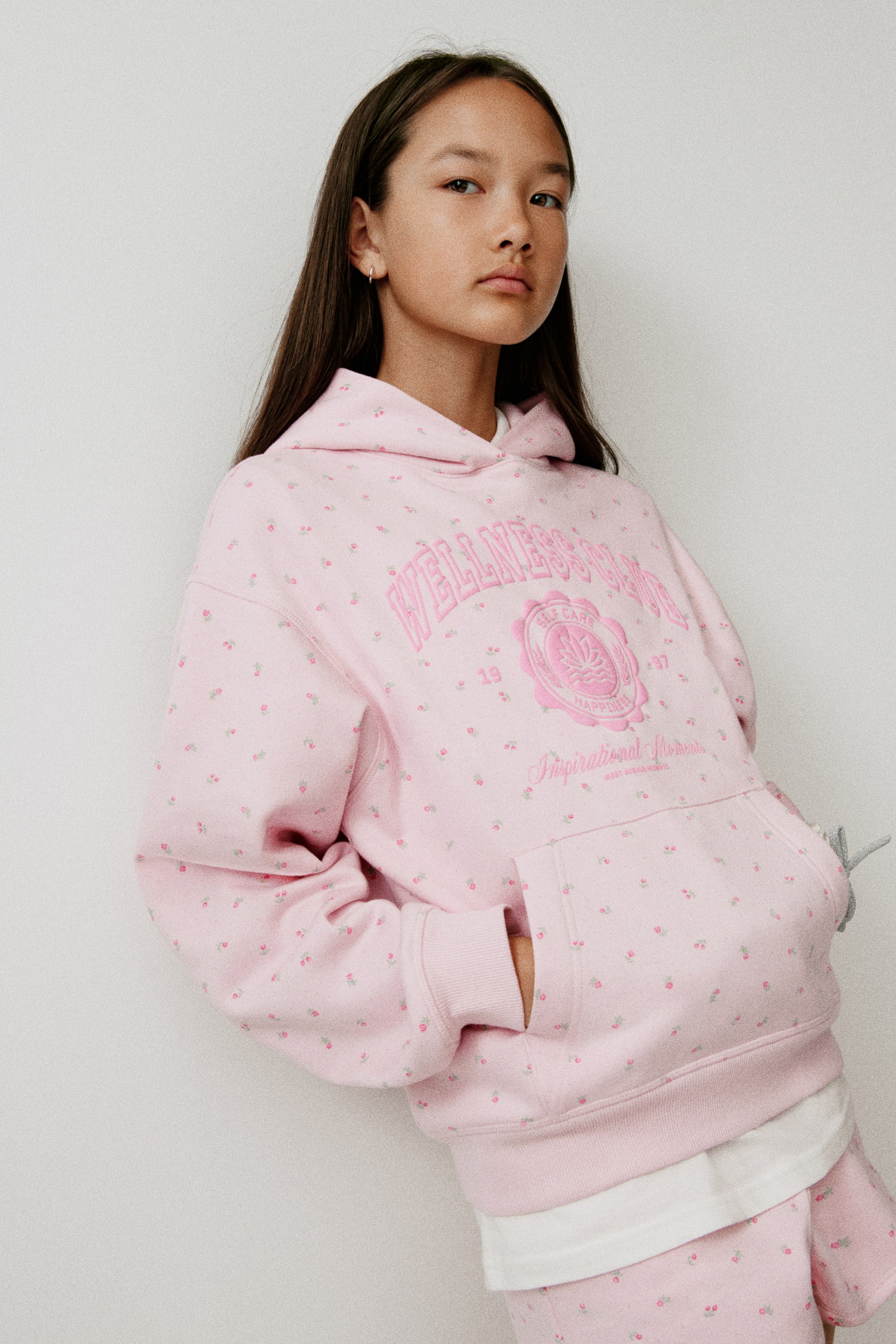 Visa större bild: En ljusrosa oversized hoodie visar ett övergripande miniblommigt tryck och ett centralt rosa grafiskt emblem med texten "WELLNESS CLUB". Hoodien har nedhasade axlar, en känguruficka framtill och breda ribbade muddar vid ärmsluten och fållen, och bärs över en vit skjorta och matchande shorts med tryck.