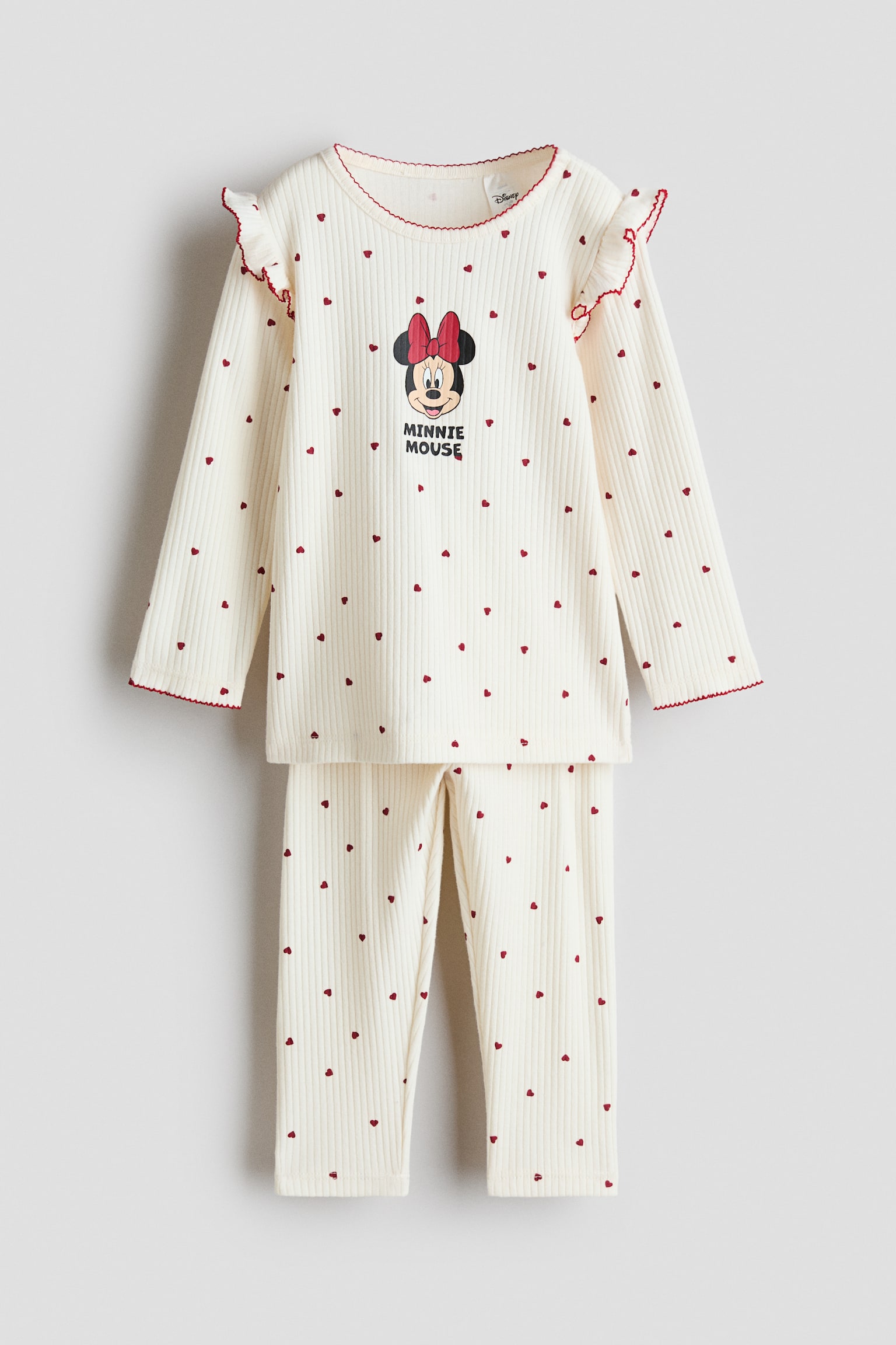 Questo pigiama color crema con motivo a coste, mostrato frontalmente, presenta un motivo a cuori rossi all-over su un top a maniche lunghe e pantaloni coordinati. Il top mostra sul petto una grafica centrale di Minnie Mouse, dettagli a volant sulle spalle e cuciture rosse con bordo ondulato lungo lo scollo e i polsini.