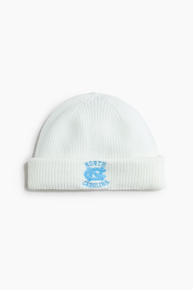Un bonnet blanc en maille cÃ´telÃ©e est prÃ©sentÃ© de face, avec un large revers repliÃ©. Le centre du revers affiche un logo brodÃ© bleu clair avec le texte Â« NORTH CAROLINA Â» en arc de cercle autour dâun symbole Â« NC Â» entrelacÃ©. - 1