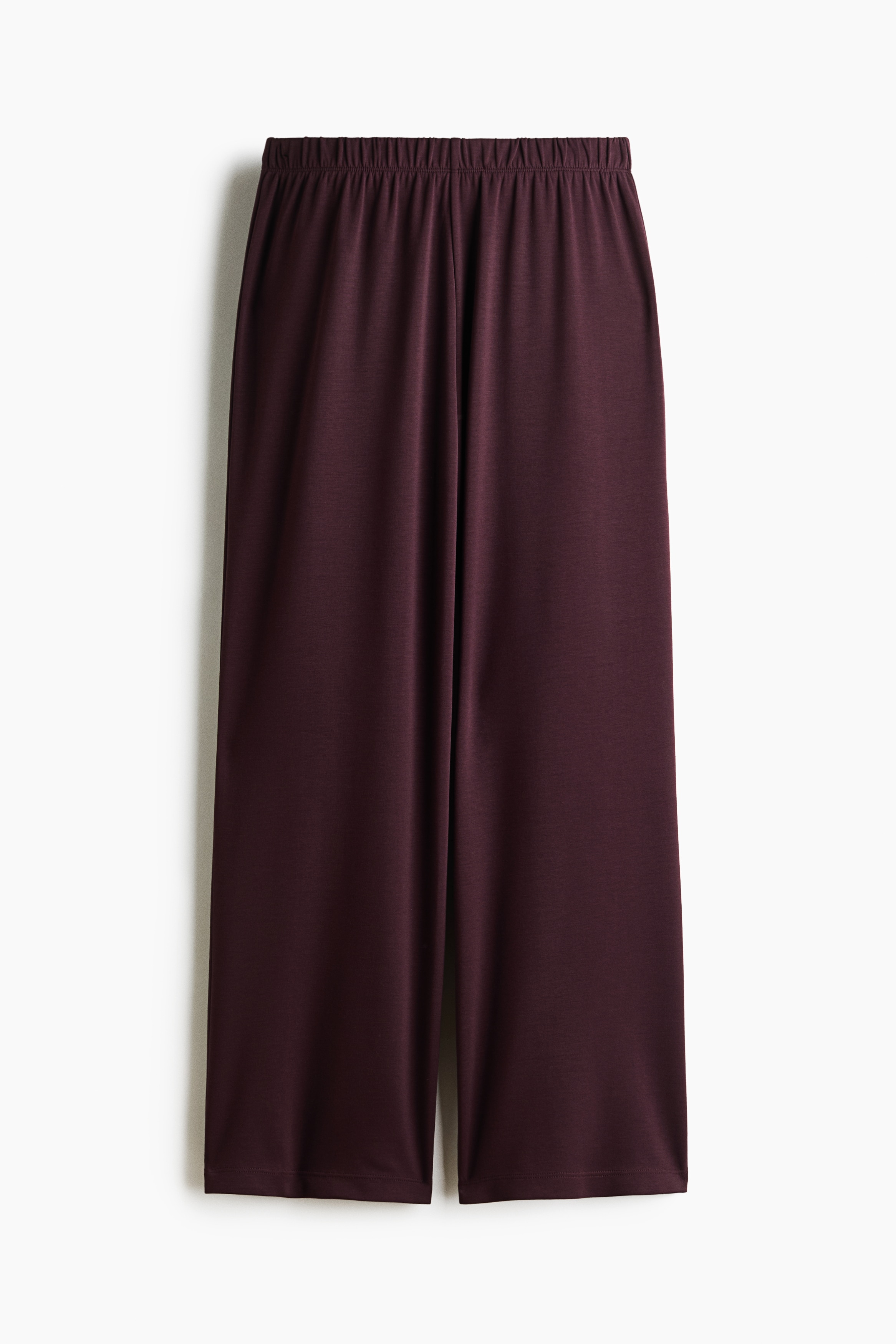 Ein bordeauxfarbenes, aufeinander abgestimmtes Set aus Half-Zip-Pullover und Hose mit weitem Bein wird von dem Model getragen. Das Model steht in einer seitlichen Profilansicht, blickt nach unten und ruht mit einer Hand nahe dem elastischen Bund der Hose, wÃ¤hrend es den Saum des locker geschnittenen Oberteils leicht anhebt.