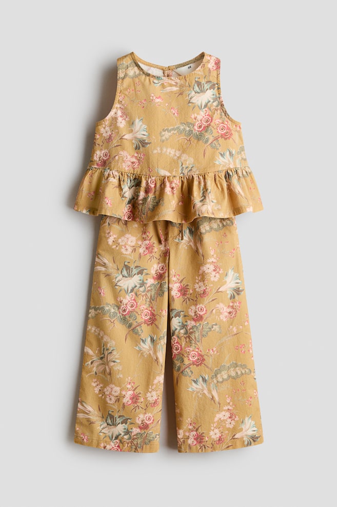This two-piece matching set in a mustard yellow floral print showcases a front view of a sleeveless top with a ruffled peplum hem and coordinating wide-leg trousers. Both pieces feature an all-over pattern of pink and white flowers with green foliage. - 1