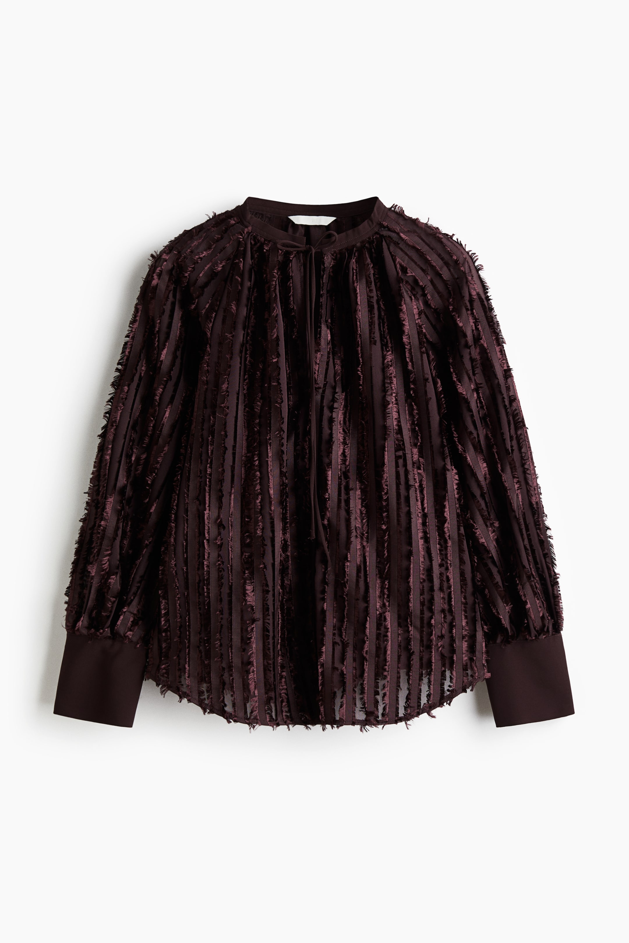 Ver imagen más grande: Una blusa de color marrón oscuro orientada hacia el frente presenta un cuello de banda con un detalle de lazo en el cuello. La prenda muestra tiras verticales de tela deshilachada a lo largo de su cuerpo y mangas, que están recogidas en los hombros y terminan en puños anchos y estructurados, con el dobladillo curvándose ligeramente.