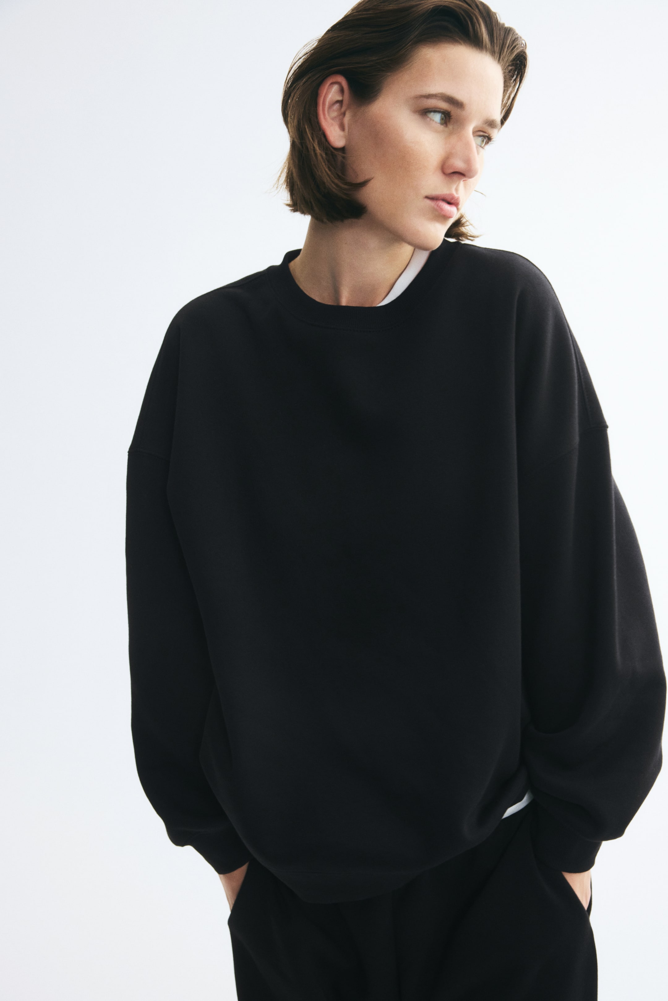 Agrandir l'image: Un sweat-shirt noir oversize à col rond, épaules tombantes et poignets côtelés est porté par le mannequin. Le sweat-shirt est superposé à un t-shirt blanc et associé à un pantalon noir assorti.