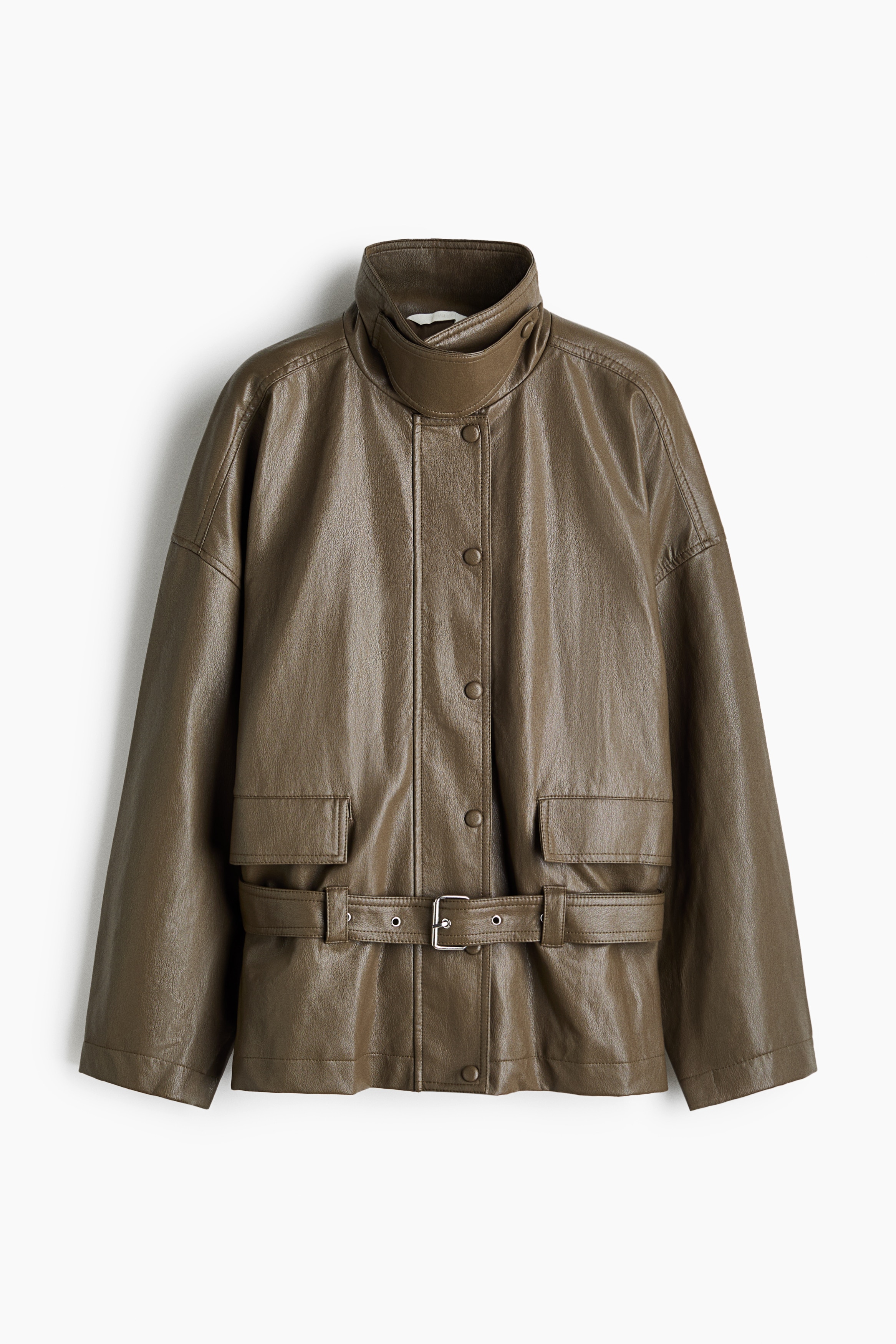 La modelo lleva una chaqueta de corte holgado en tejido recubierto verde oliva, con hombros caÃ­dos, grandes bolsillos delanteros con solapa y un cinturÃ³n ajustable a juego ceÃ±ido a la cintura. La chaqueta se lleva superpuesta sobre una camisa de cuadro gris y un jersey negro de cuello vuelto, combinada con pantalones negros.