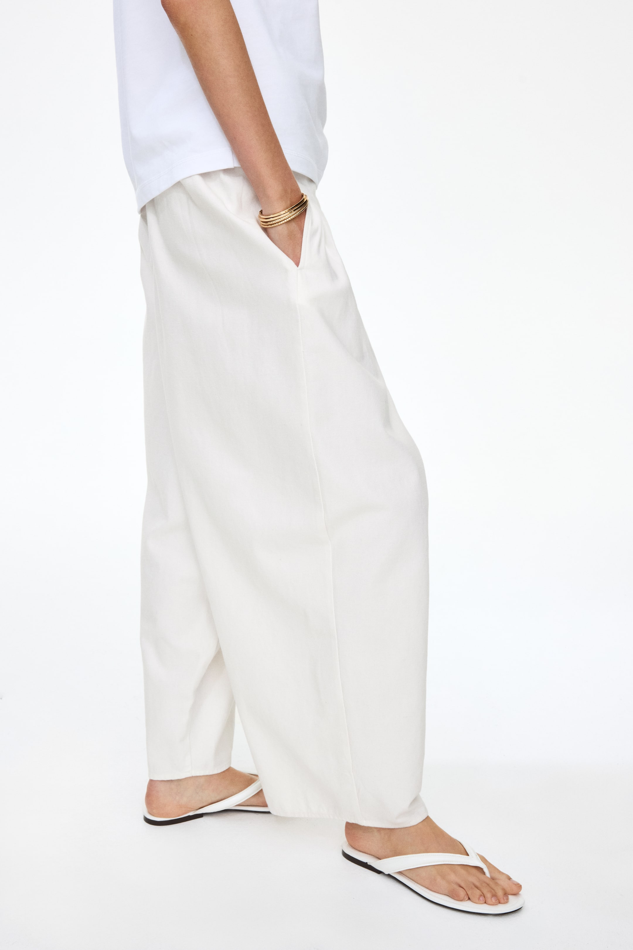 Visualizza immagine più grande: I pantaloni in tela di cotone bianco a lunghezza caviglia e vestibilità ampia sono indossati dalla modella. Presentano una vita regolare, gambe a barile voluminose e tasche laterali diagonali, con la mano della modella che riposa in una di esse. La modella abbina i pantaloni a una t-shirt bianca e infradito bianche.