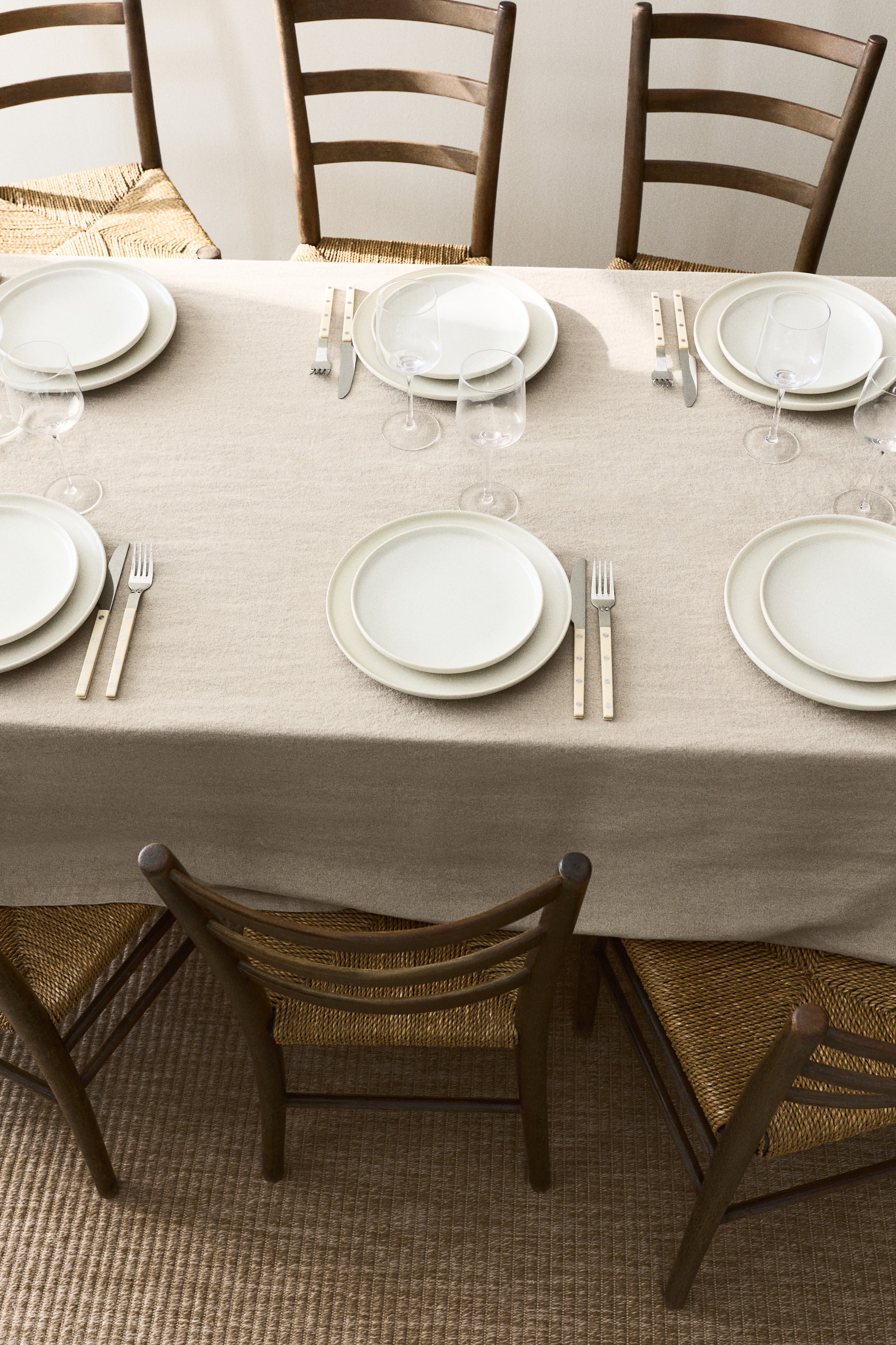 Voir une image plus grande: Les couverts sont disposés sur une nappe beige clair, vus d'en haut. Chaque couvert comprend deux assiettes rondes blanc cassé, une fourchette et un couteau avec des manches de couleur claire de chaque côté, et des verres à pied transparents à droite.