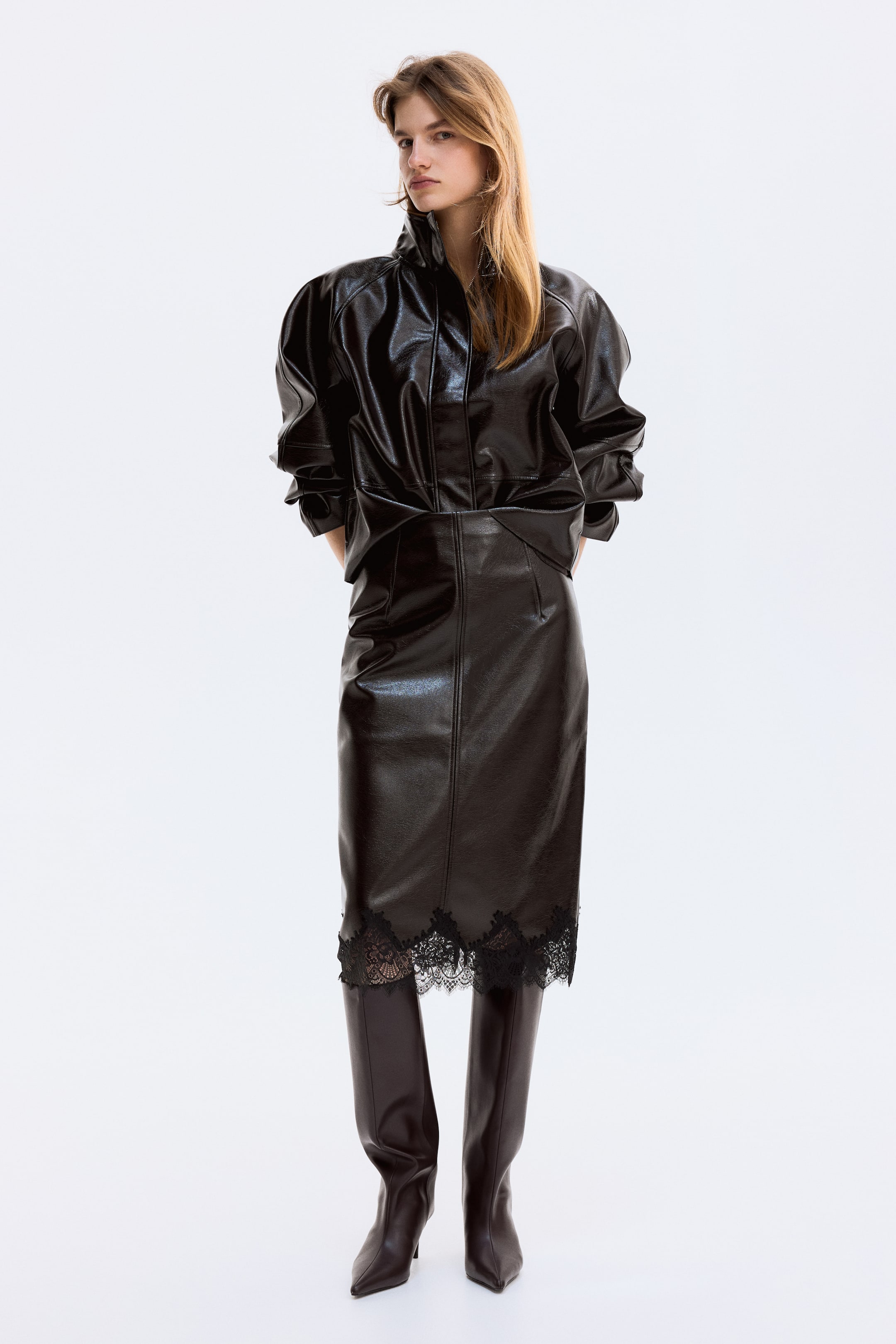 Ver imagen más grande: Un conjunto oscuro de aspecto cuero brillante se presenta, con una blusa estilo blusón de frente con cuello alto y mangas voluminosas, combinado con una falda lápiz ajustada. La falda se extiende por debajo de la rodilla y presenta un ribete de encaje negro en el dobladillo, mientras que unas botas oscuras de punta afilada y hasta la rodilla completan el look.