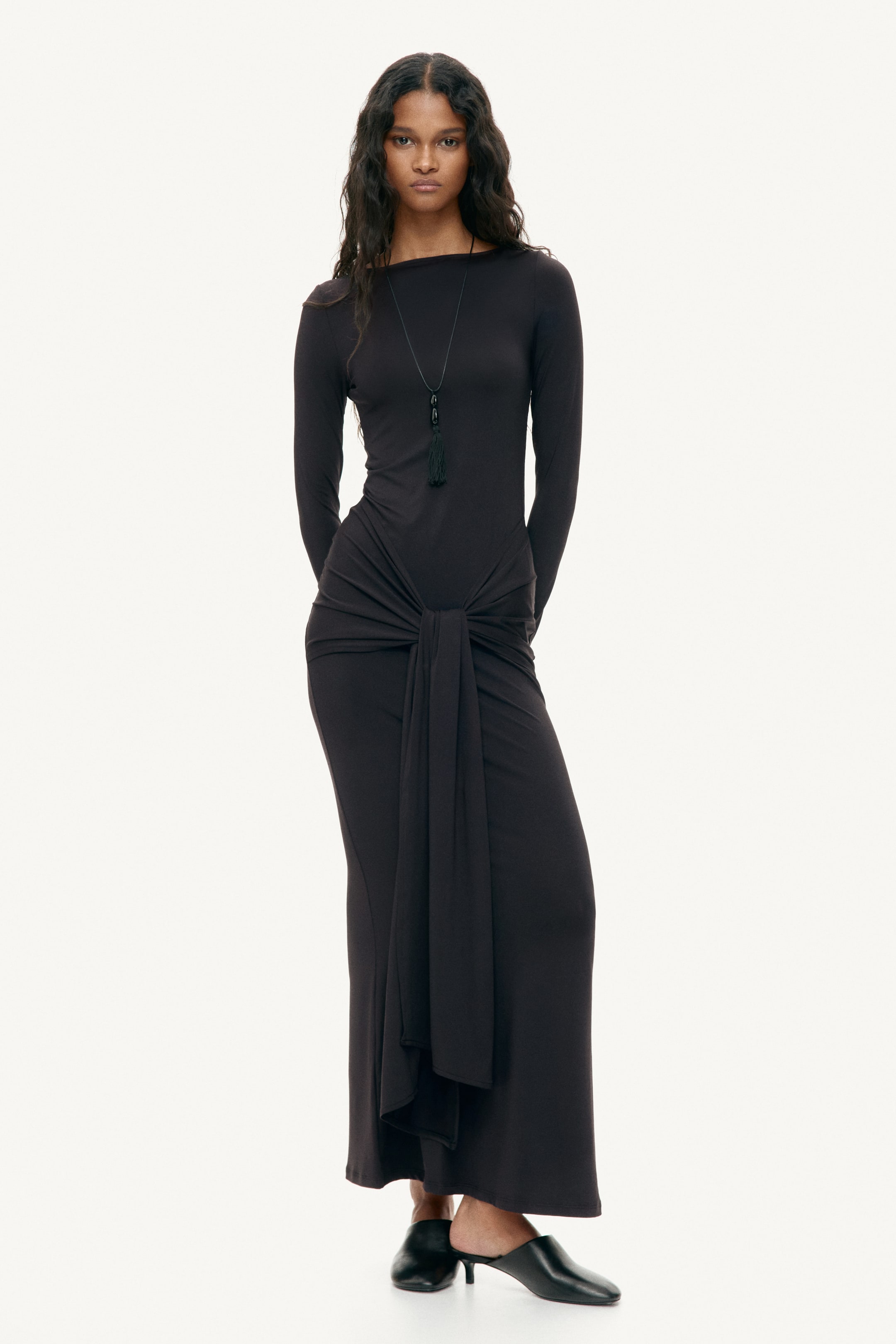 View larger image: The model is wearing a long, fitted black dress made of stretch jersey, showcasing a boat neckline and long sleeves. A wide section at the hips is tied at the front, creating a draped effect. The model complements the dress with a black necklace and black mules.