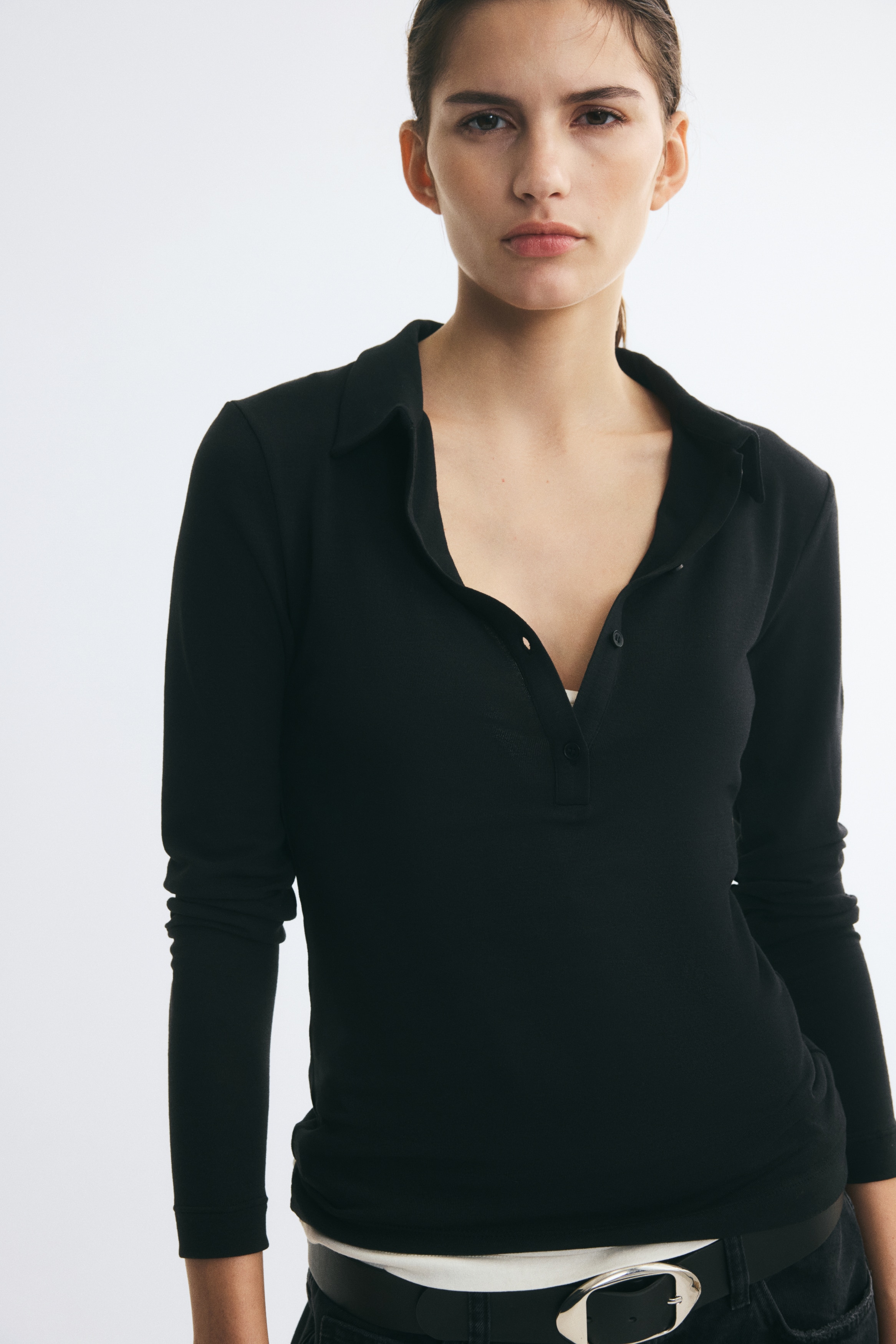 Un tricou polo negru, lung Èi cambrat, din jerseu elastic, este prezentat din faÈÄ, avÃ¢nd un guler clasic, o deschidere verticalÄ cu patru nasturi Èi mÃ¢neci extra-lungi.