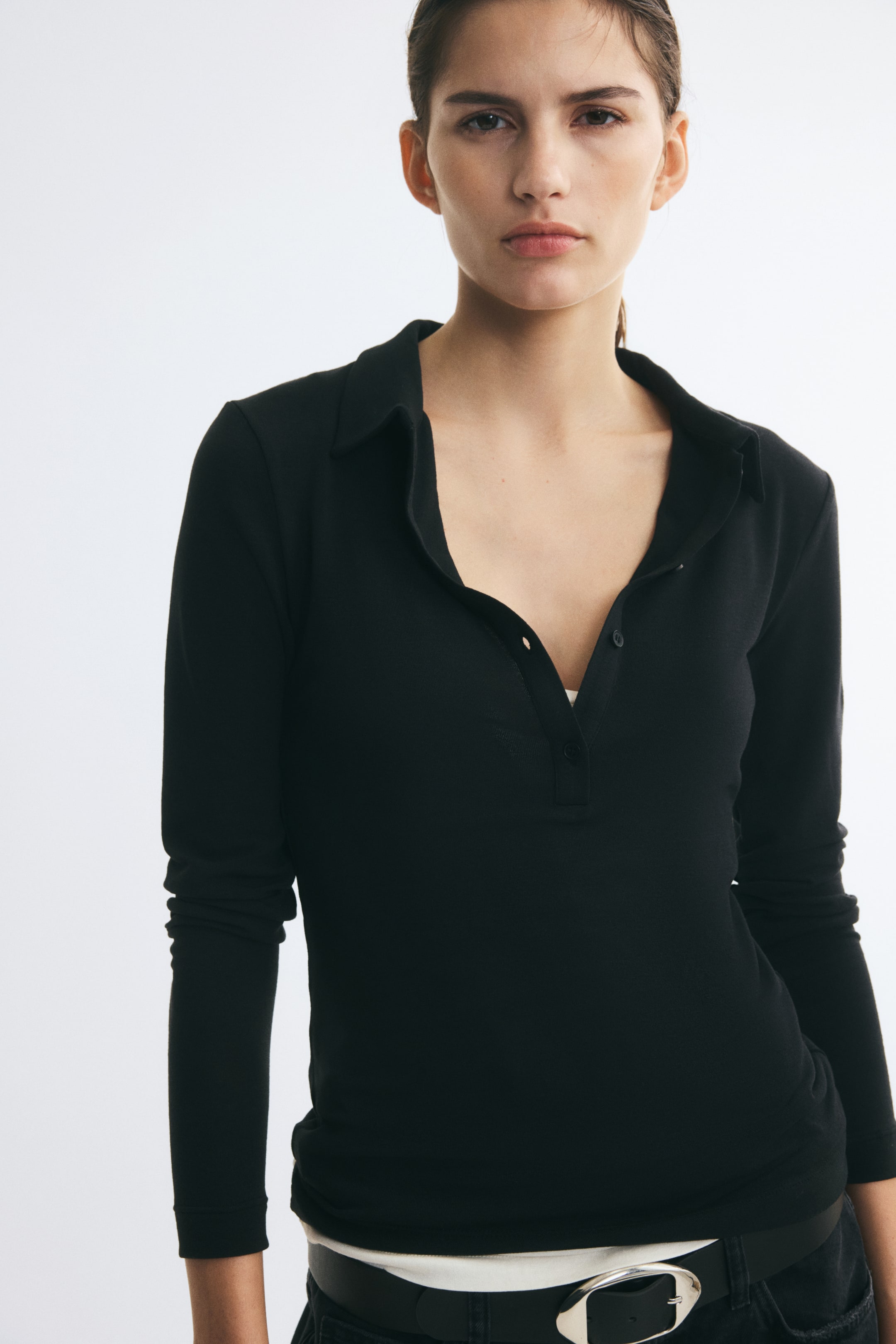 Agrandir l'image: Un polo noir long et ajusté en jersey extensible est porté par le mannequin, avec un col, une patte de boutonnage ouverte et des manches longues. Le polo est porté sur un haut blanc et associé à un pantalon noir ainsi qu'à une ceinture avec boucle argentée.