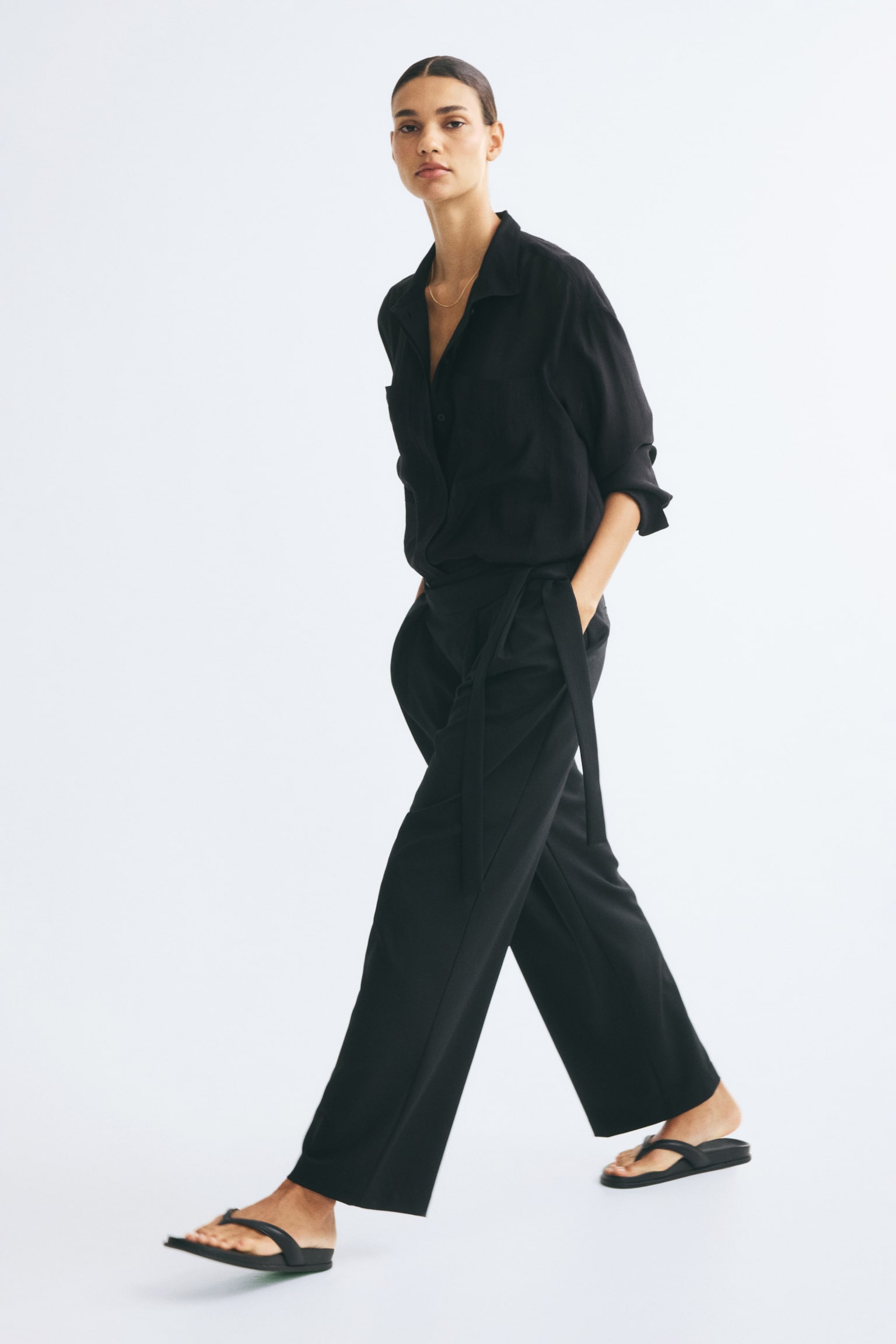 Black loose-fit trousers featuring a high-waisted wrap front with hanging side ties, front pleats, and wide legs are being worn by the model. The model is walking while wearing the trousers, which are styled with a tucked-in black long-sleeve shirt and black sandals. - 1