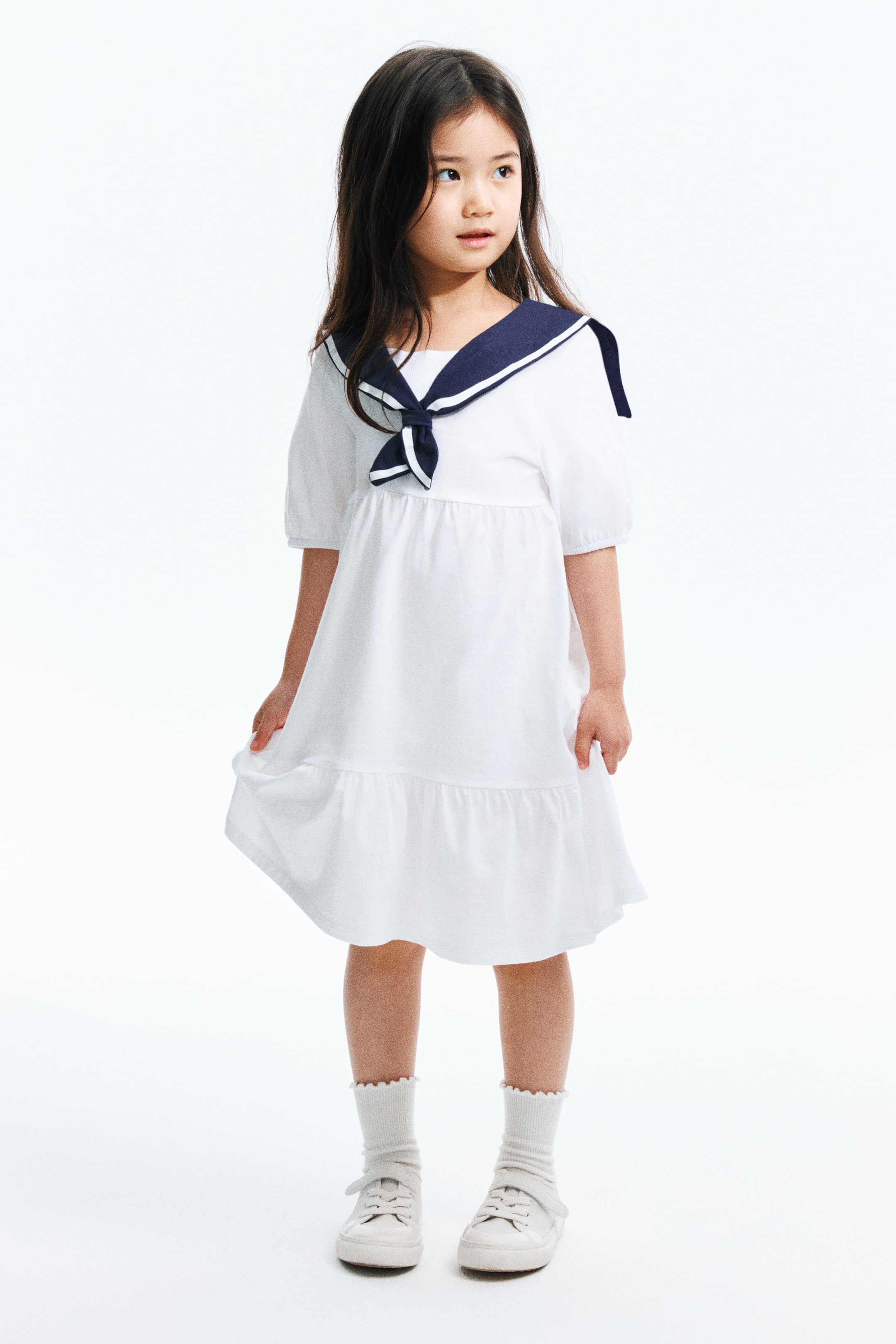 Xem ảnh lớn hơn: Một chiếc váy trắng nổi bật với cổ áo thủy thủ màu xanh navy có viền trắng, được thắt nút ở phía trước, và tay áo ngắn phồng. Phần váy xếp ly được thiết kế với một tầng bèo ở gấu, tạo dáng đầy đặn khi nhìn từ phía trước.