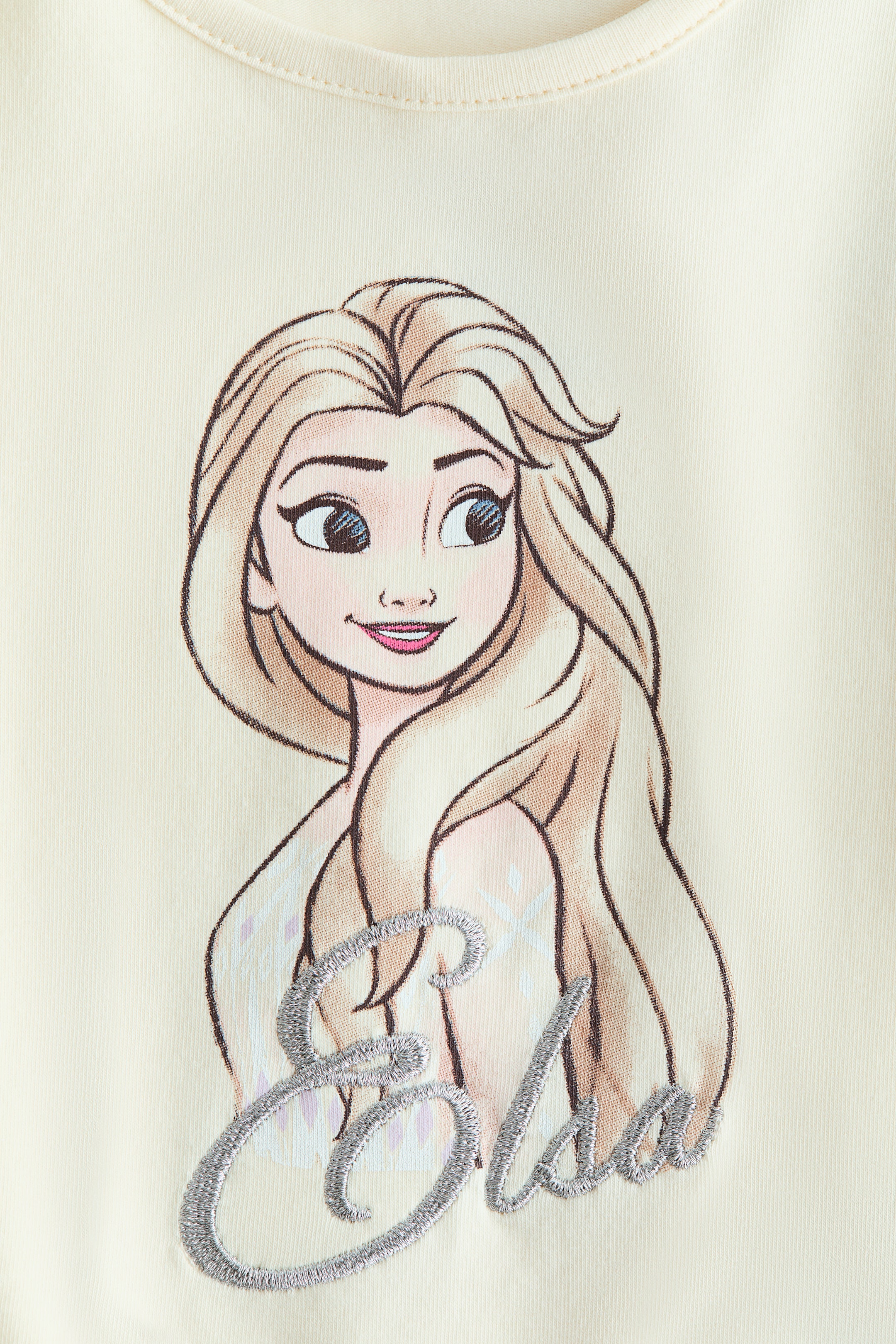 Προβολή μεγαλύτερης εικόνας: Ένα μακρυμάνικο τοπ από μαλακό βαμβακερό ζέρσεϊ φαίνεται από μπροστά, με στρογγυλή λαιμόκοψη και στενό τελείωμα. Στο στήθος του, υπάρχει ένα μοτίβο που απεικονίζει μια φιγούρα θηλυκού χαρακτήρα με ξανθά μαλλιά και μπλε μάτια, και κάτω από αυτήν, το όνομα "Elsa" είναι κεντημένο με ασημένια κυρτή γραφή.