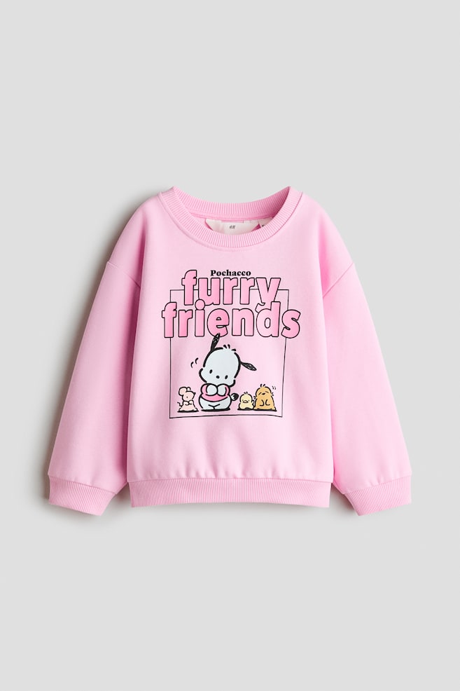 Uma sweatshirt rosa está vista de frente, com uma imagem impressa do personagem de desenho animado Pochacco e de outros pequenos animais por baixo do texto "Pochacco furry friends". A peça apresenta gola redonda canelada, ombros descaÃdos, mangas compridas com punhos canelados e bainha canelada. - 1