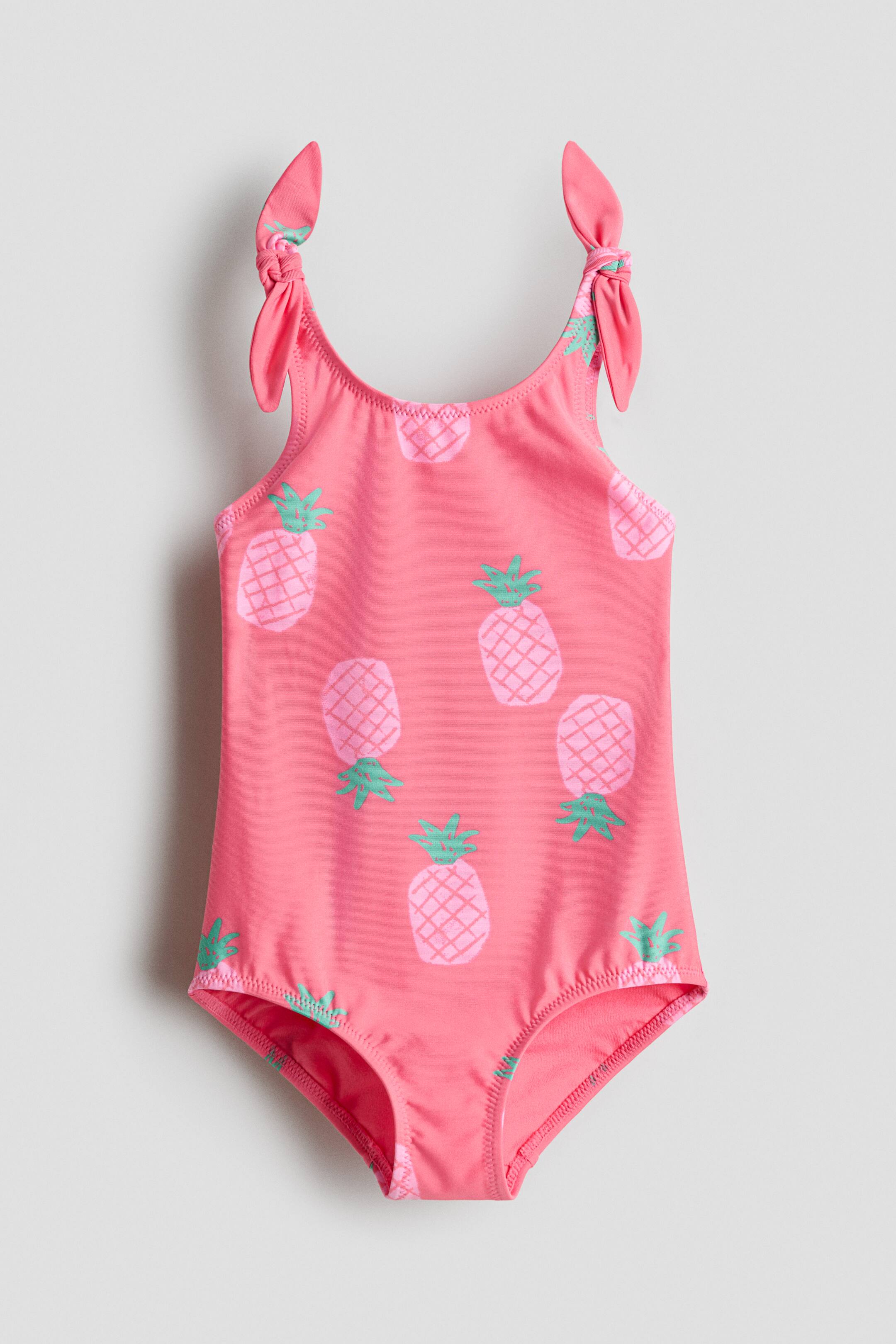 Ingrandisci l'immagine: Un costume da bagno rosa presenta una stampa di ananas su tutta la superficie in rosa più chiaro e verde, presentato FRONTALE. È progettato con spalline che formano dettagli decorativi a nodo.