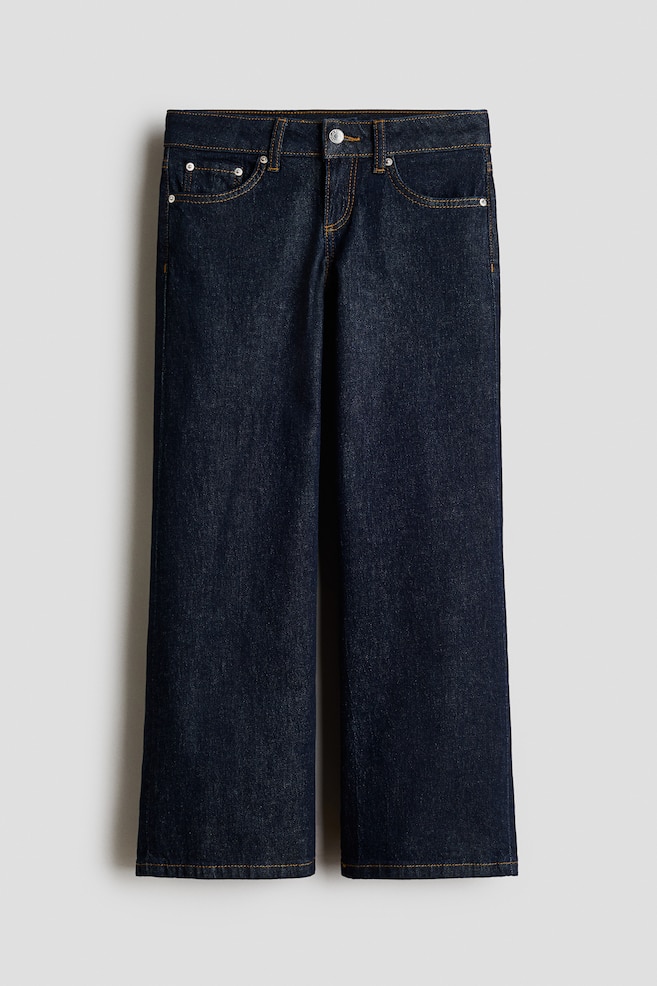 A pair of dark indigo wide-leg cotton denim jeans is hanging in a front view, displaying a classic five-pocket design with orange contrast topstitching. The jeans feature a low waist with a single metal button fastening and a zip fly. - 1