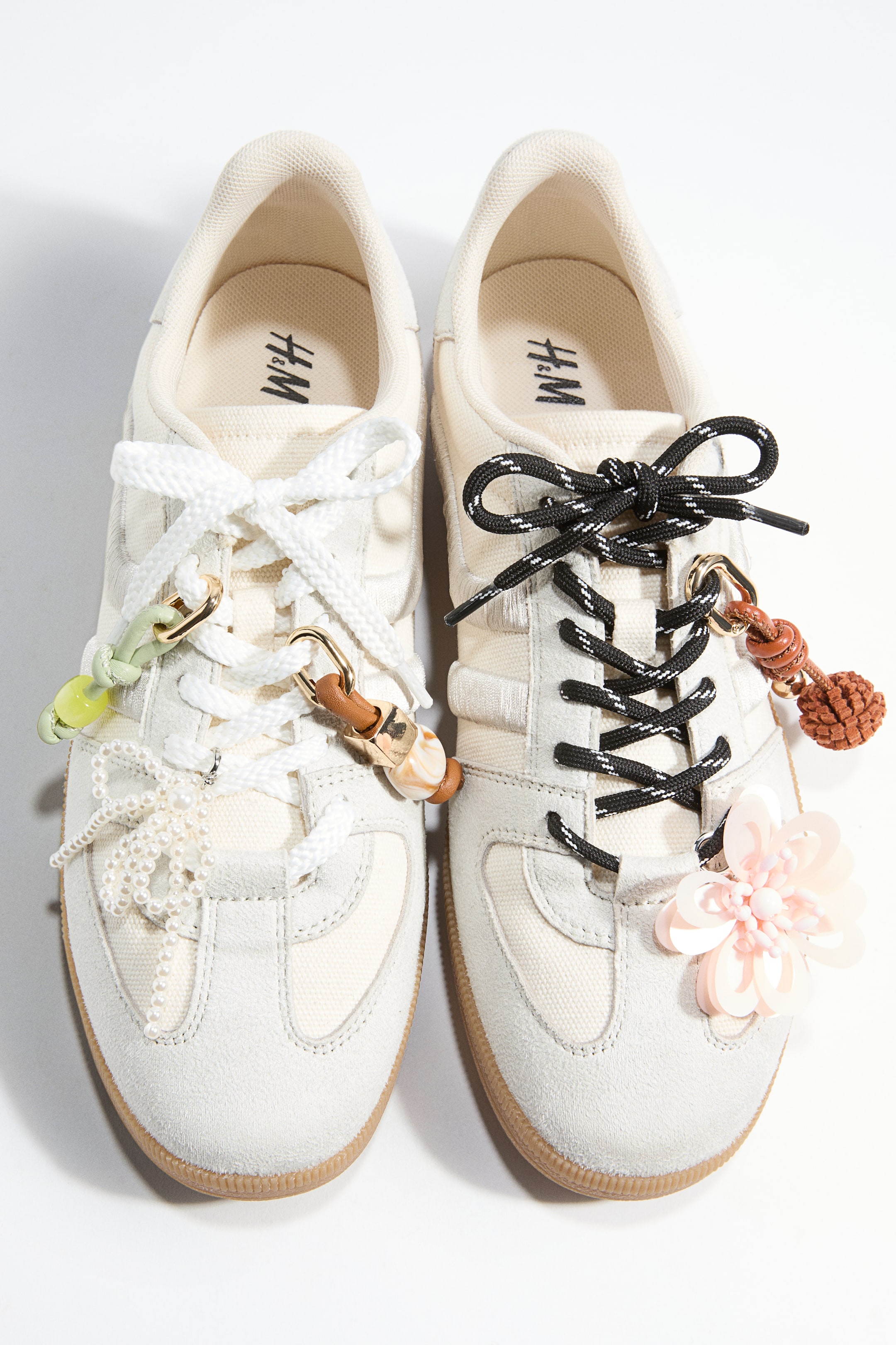 Vis større bilde: To off-white sneakers, konstruert med semsket overlegg og canvaspaneler, vises fra et topp-ned perspektiv. Den venstre sneakeren har hvite lisser pyntet med en grønn perlecharme, en brun lær- og gullcharme, og en rekke hvite perler, mens den høyre sneakeren viser frem svart- og hvitstripete lisser med en brun flettet dusk og en rosa blomsterapplikasjon.