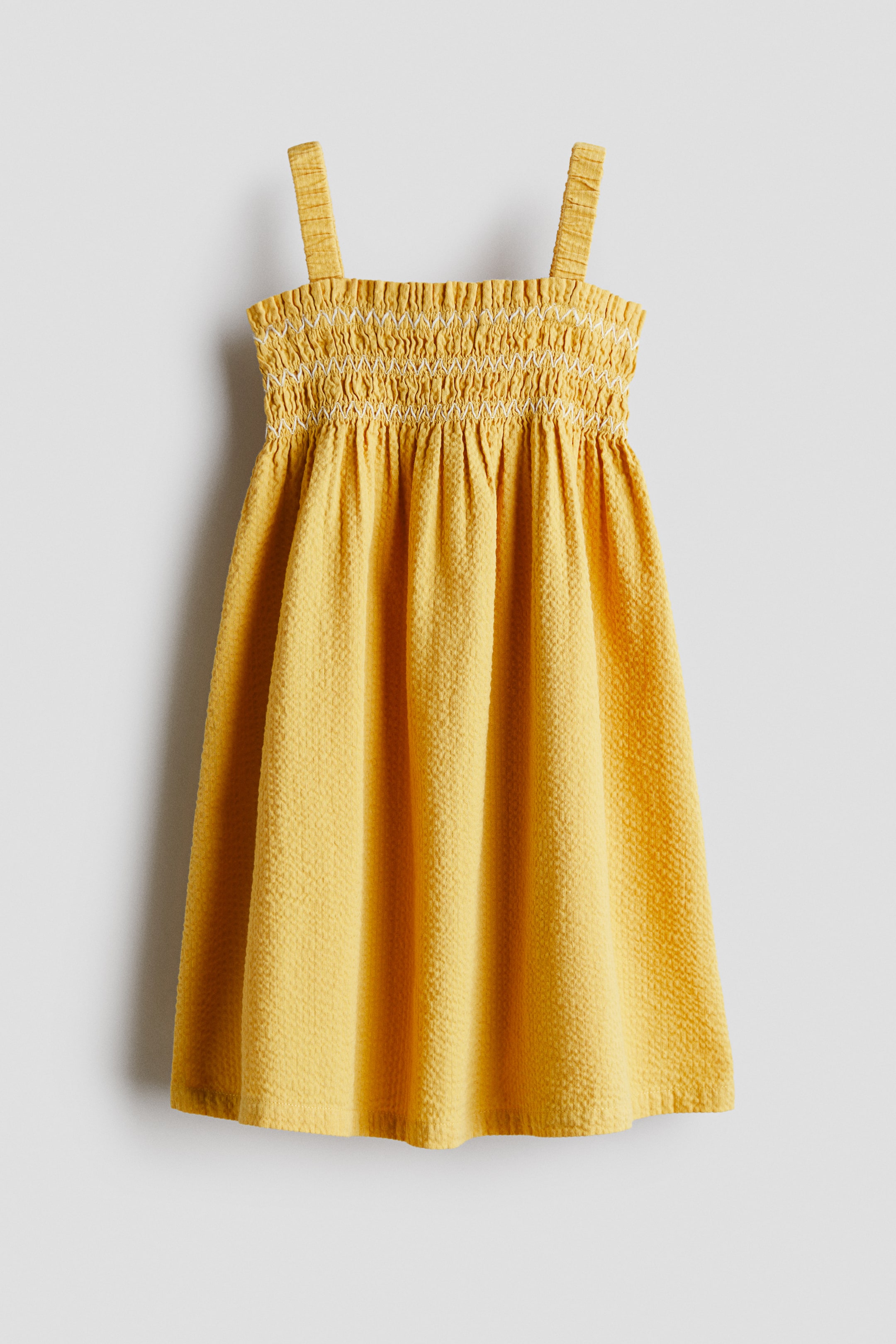 View larger image: A yellow textured dress is shown from the front, featuring a smocked bodice with decorative white zig-zag embroidery and thick shoulder straps. The dress falls into a gathered, flared skirt made from crinkled seersucker fabric.