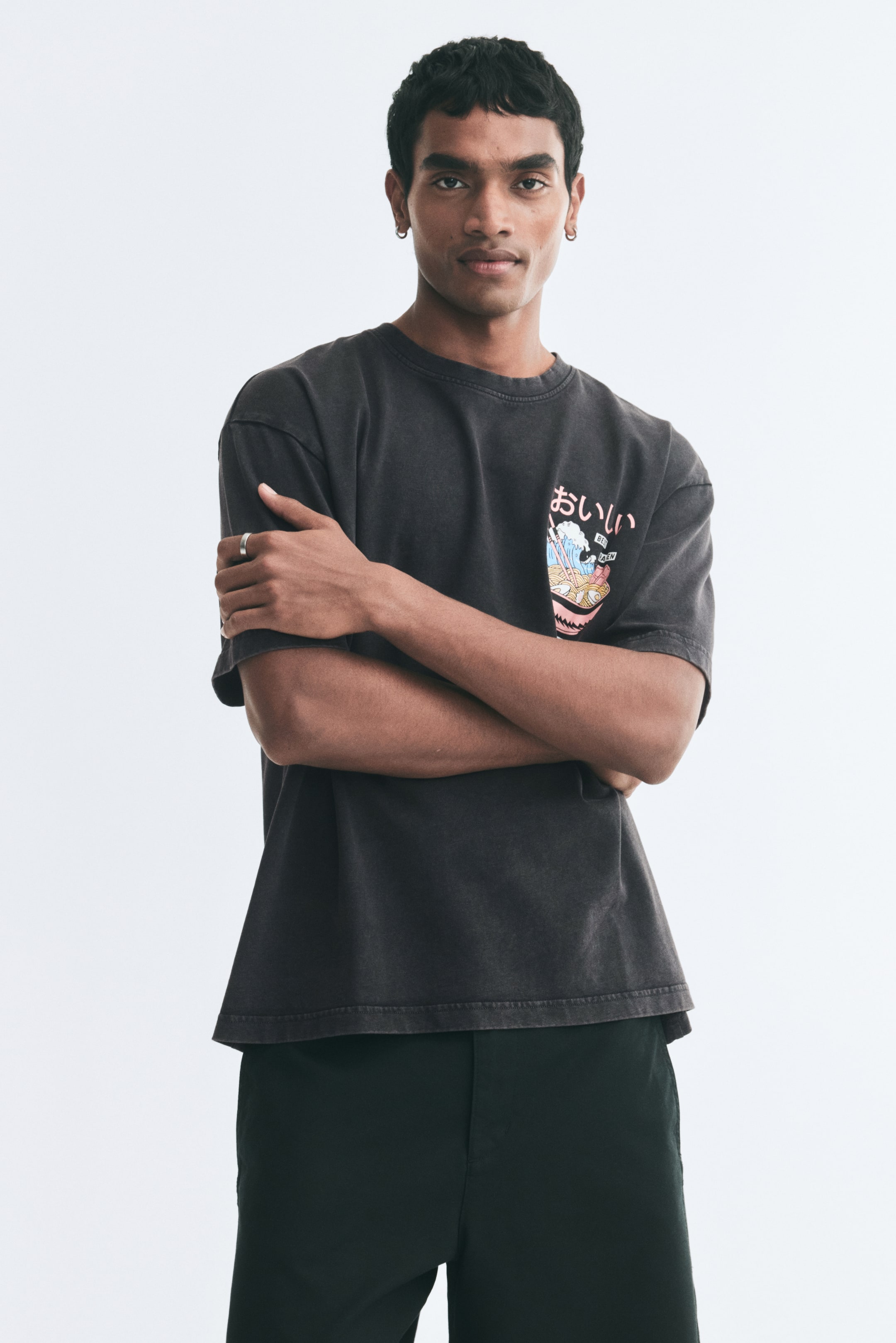 View larger image: A dark grey, relaxed-fit cotton T-shirt is being worn by the model, who is standing with arms crossed. The T-shirt features a round, ribbed neckline, dropped shoulders, and a colourful graphic design of a ramen bowl with Japanese characters printed on the left chest.