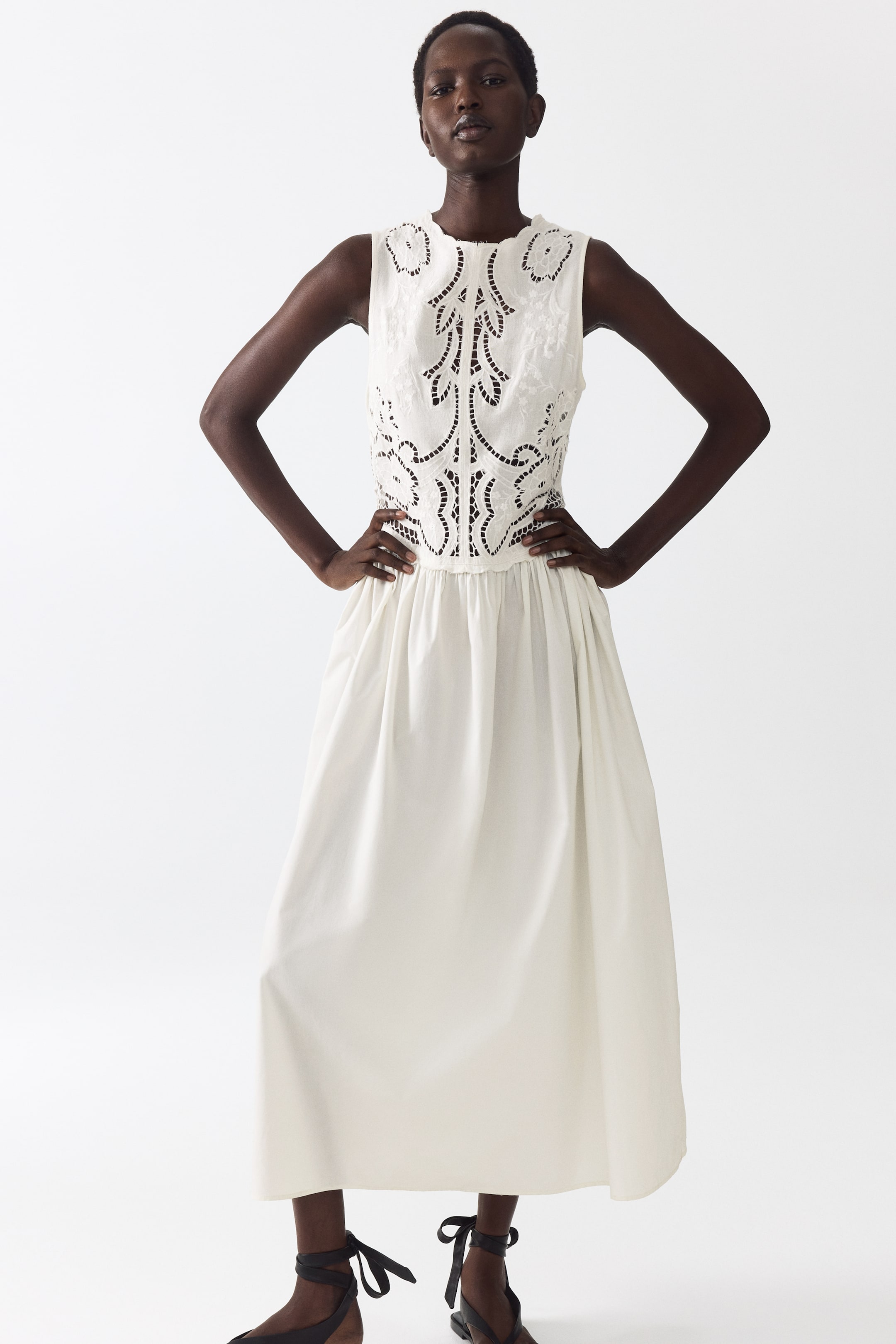 Agrandir l'image: Une robe midi sans manches blanc cassé est présentée de face, mettant en avant un corsage brodé avec des découpes complexes et un col rond. Une taille froncée sépare le corsage d'une jupe ample et fluide qui s'étend jusqu'à mi-mollet.