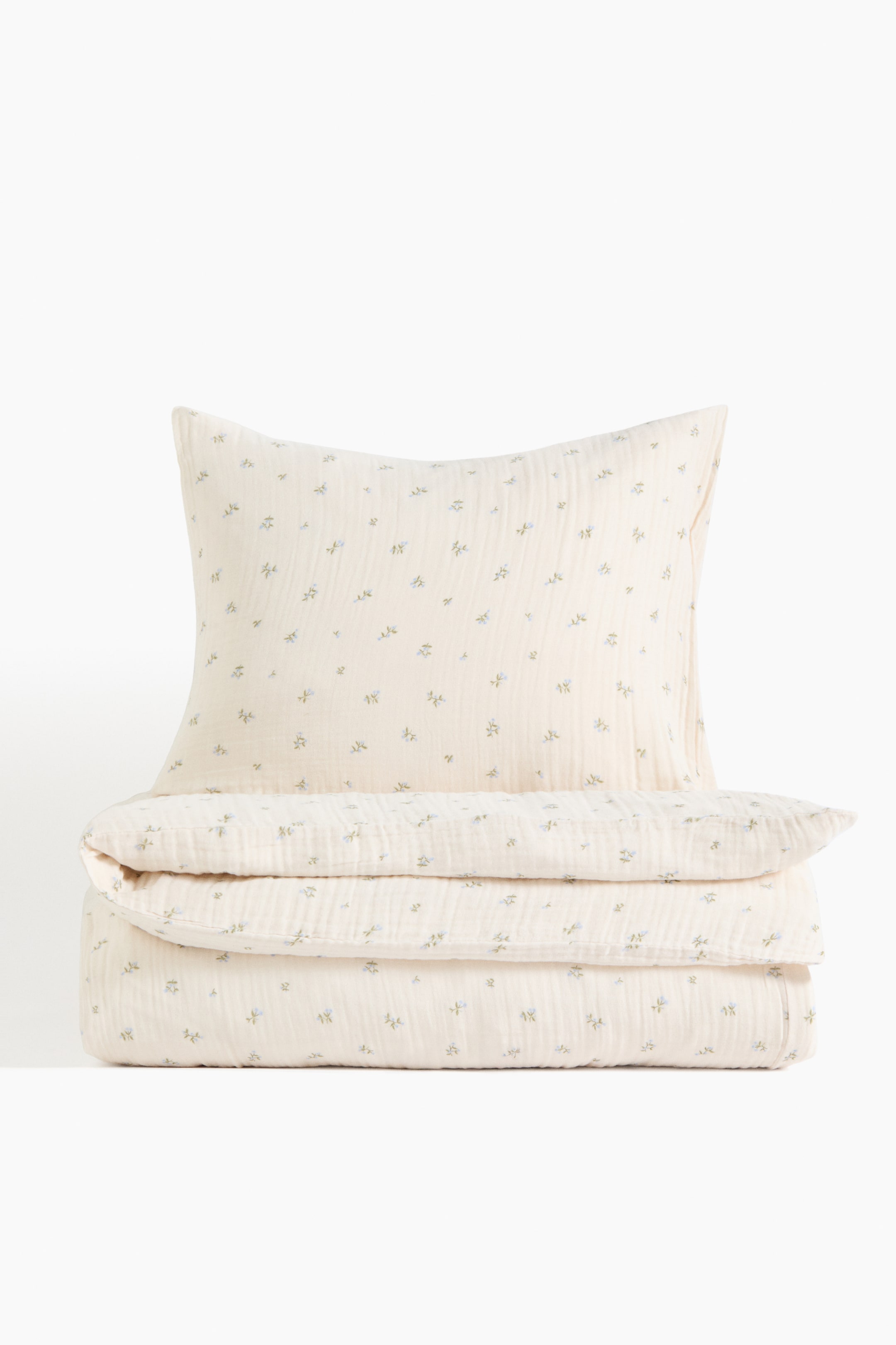 Größeres Bild ansehen: Ein cremefarbenes Bettbezug-Set aus weicher Baumwollmusselin mit einem durchgehenden Druck kleiner blauer Blumenmotive. Das Set präsentiert einen Kissenbezug, der aufrecht hinter einem ordentlich gefalteten Bettbezug steht.