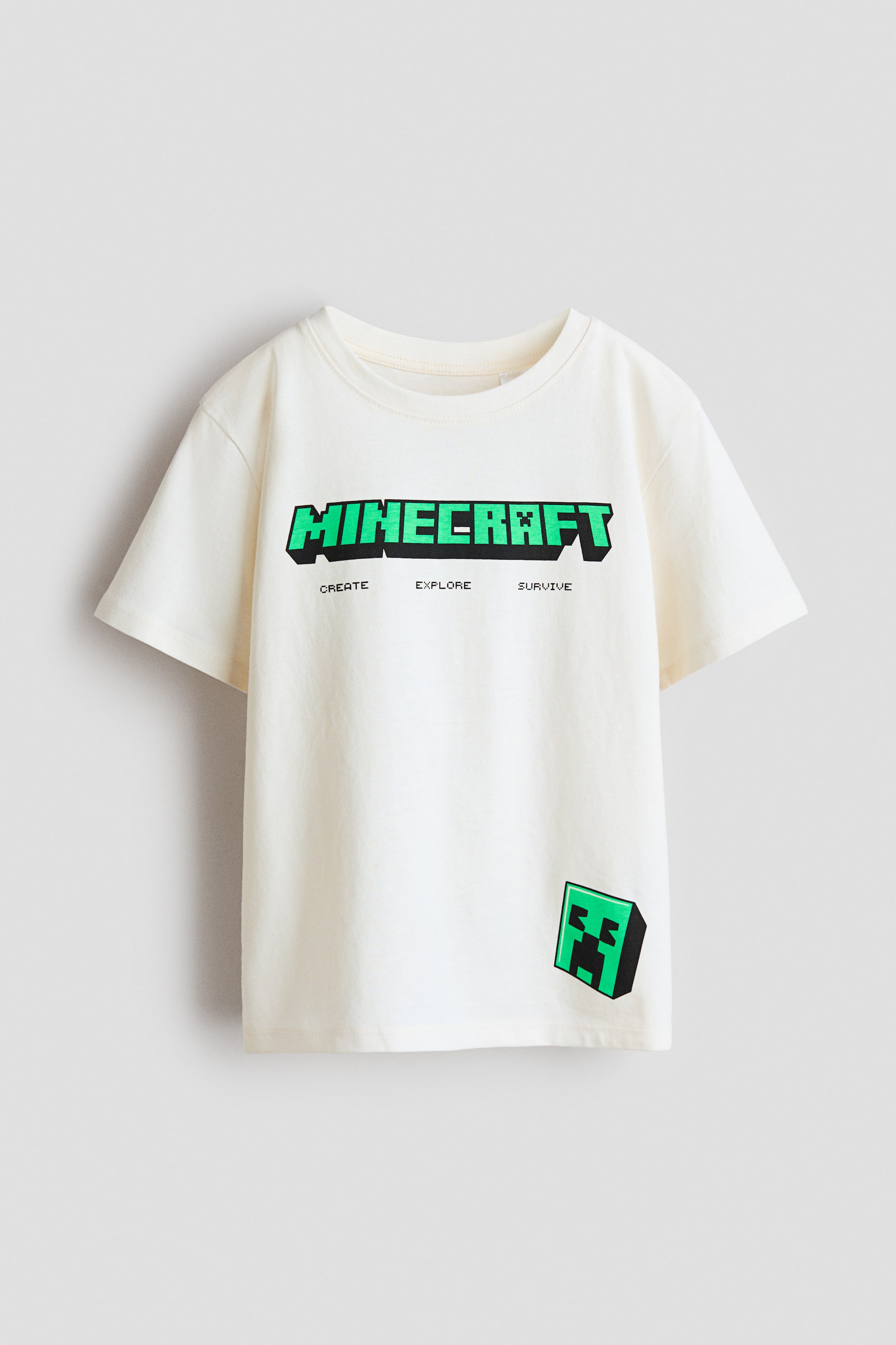 View larger image: A t-shirt berwarna krim dipersembahkan rata, menghadap depan. Ia menampilkan leher bulat berusuk, lengan pendek, dan logo "MINECRAFT" berwarna hijau dalam bentuk piksel di bahagian dada. Di bawah logo, perkataan "CREATE EXPLORE SURVIVE" dicetak, dan wajah Creeper berbentuk piksel hijau dipaparkan di bahagian depan kanan bawah.