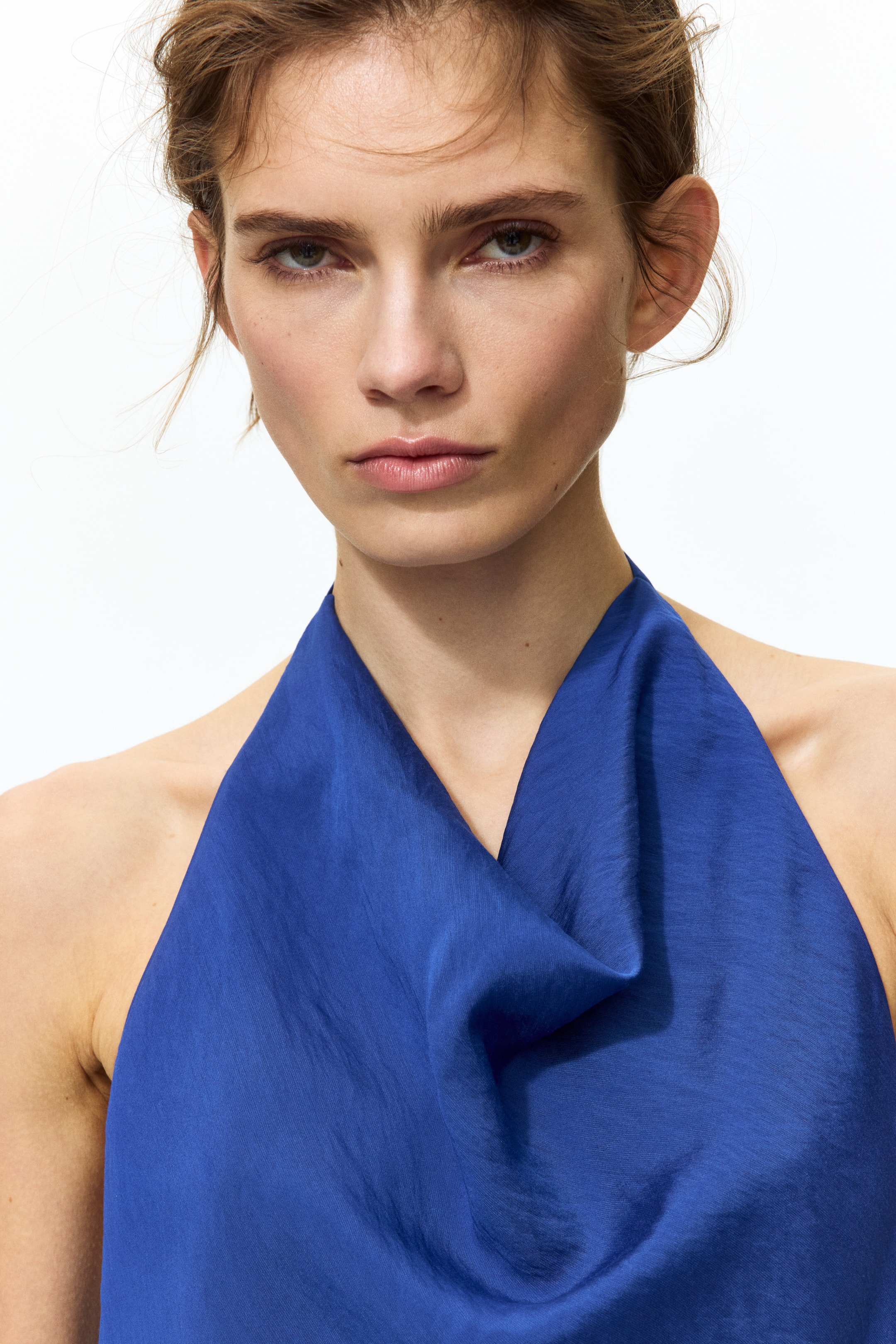 View larger image: A deep blue halter top features a draped cowl neckline with soft folds. This sleeveless piece showcases a fabric with a subtle sheen on the front.