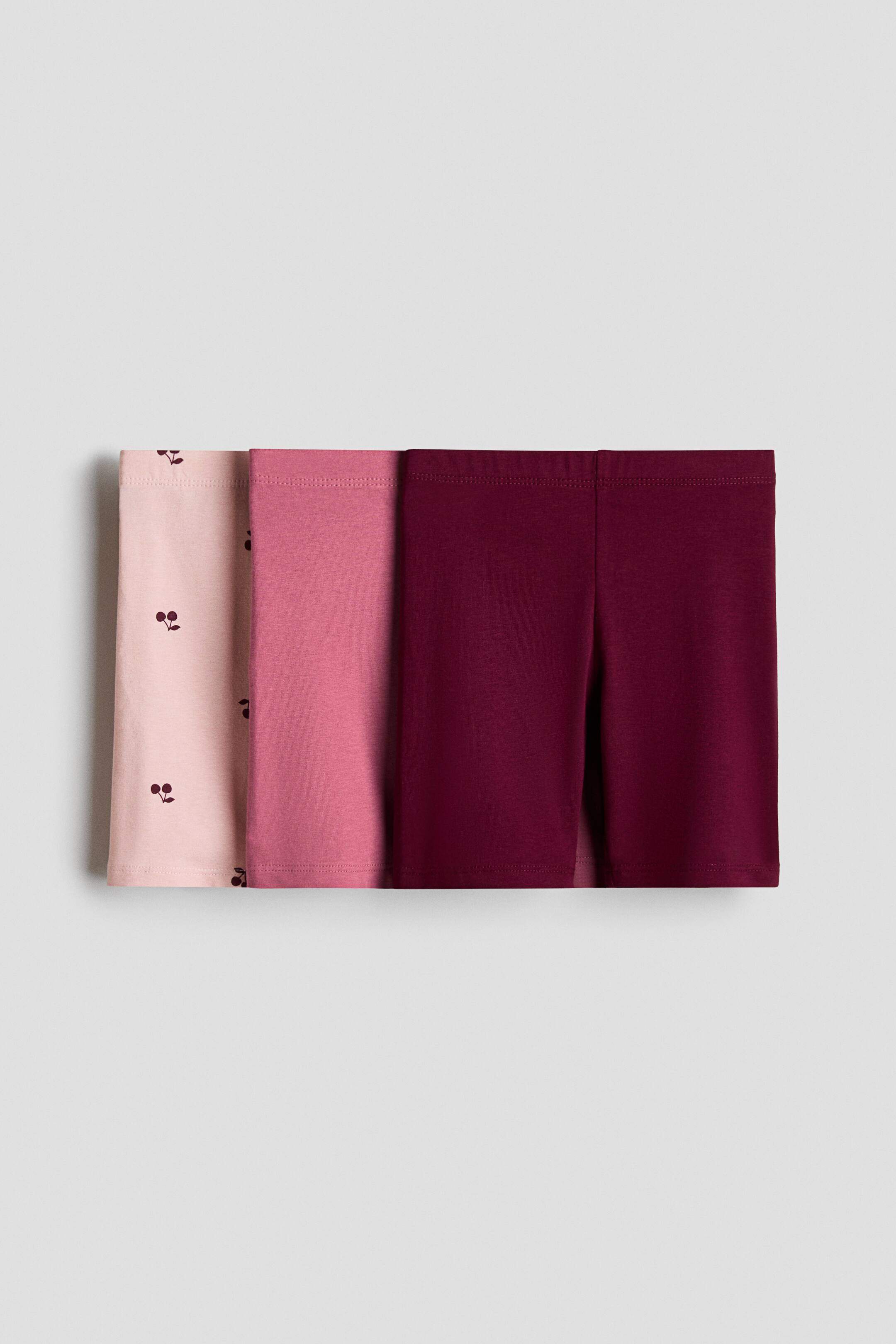 View larger image: Three pairs of cycling shorts are shown, arranged side by side with a slight overlap, featuring elastic waistbands and a soft jersey texture. The shorts come in light pink with a cherry pattern, solid pink, and solid burgundy.