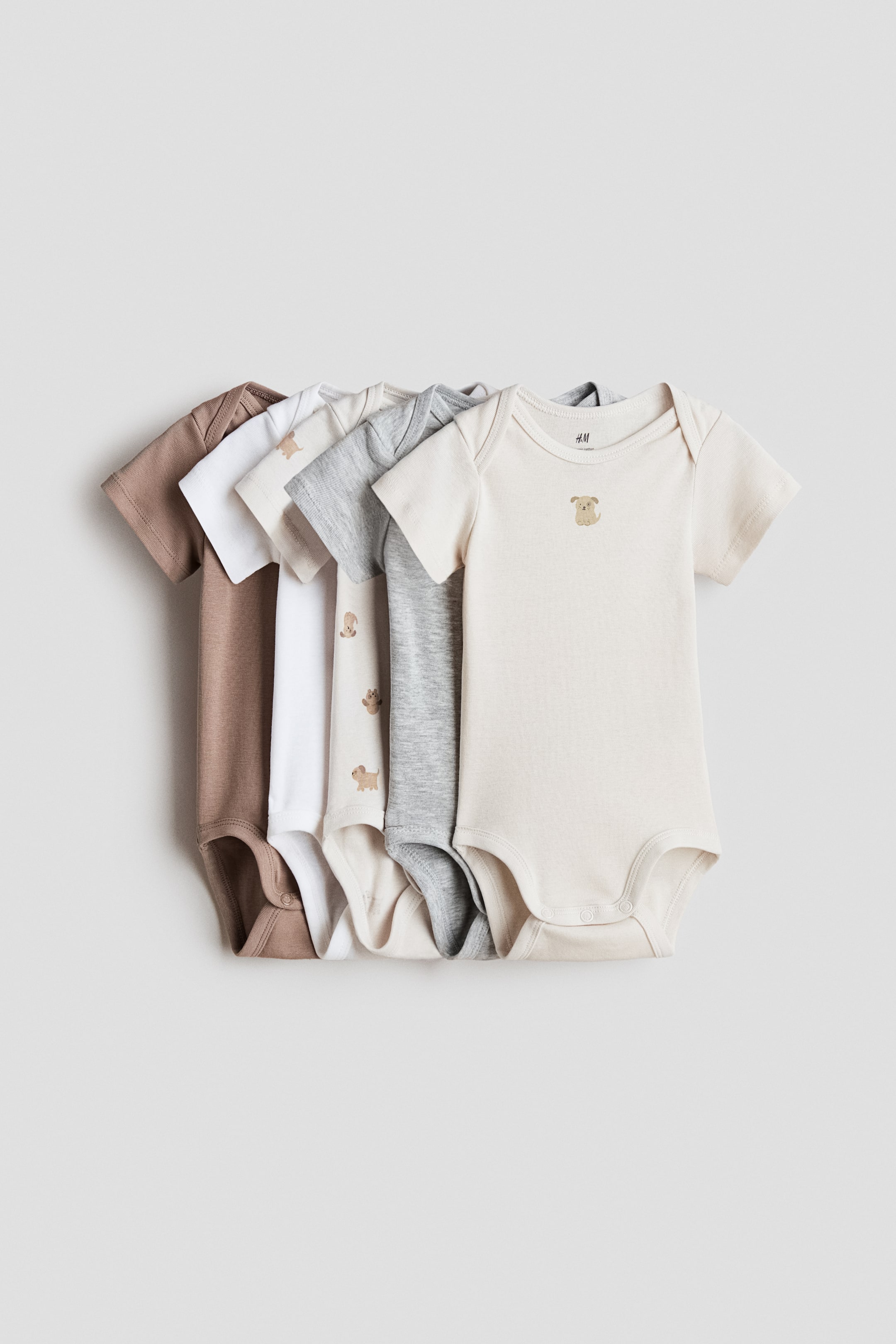 Zobrazit větší obrázek: Sada pěti bodýček s krátkými rukávy ze supersoft cotton jersey je uspořádána čelem dopředu (FRONT), naskládaná a rozvěšená do vějíře v odstínech hnědé, bílé, světle béžové, šedé a krémové. Každé bodýčko má překládaná ramena (lapped shoulders) a press-studs v rozkroku; krémové bodýčko nese na hrudi malého vyšívaného psa a světle béžové zobrazuje několik vyšívaných psů.