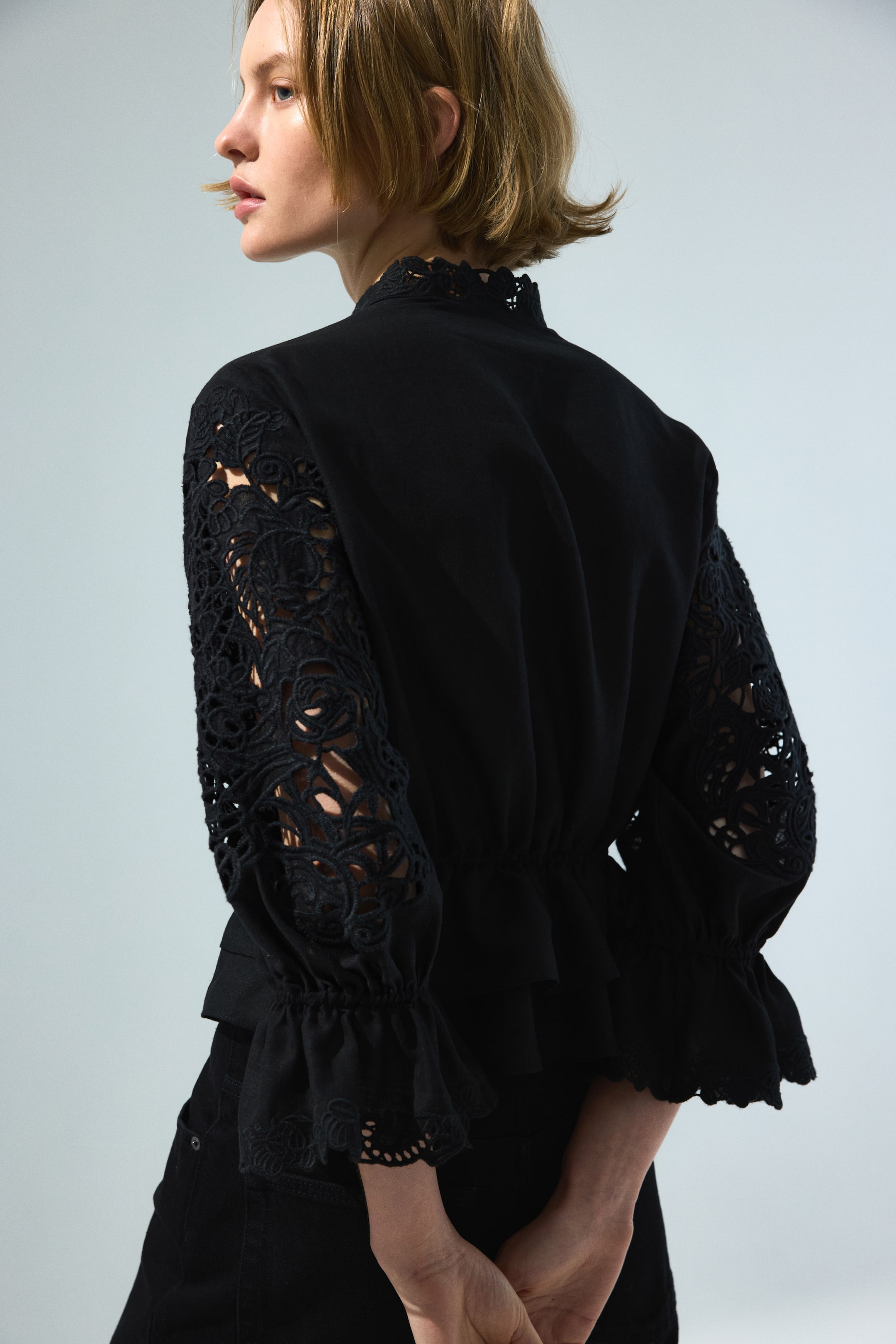 View larger image: A black blouse is showcasing a back-side view, featuring a high scalloped collar. Its long sleeves display intricate black lace cutouts on the upper arms and gather into ruffled cuffs, while the waist cinches to form a subtle peplum.