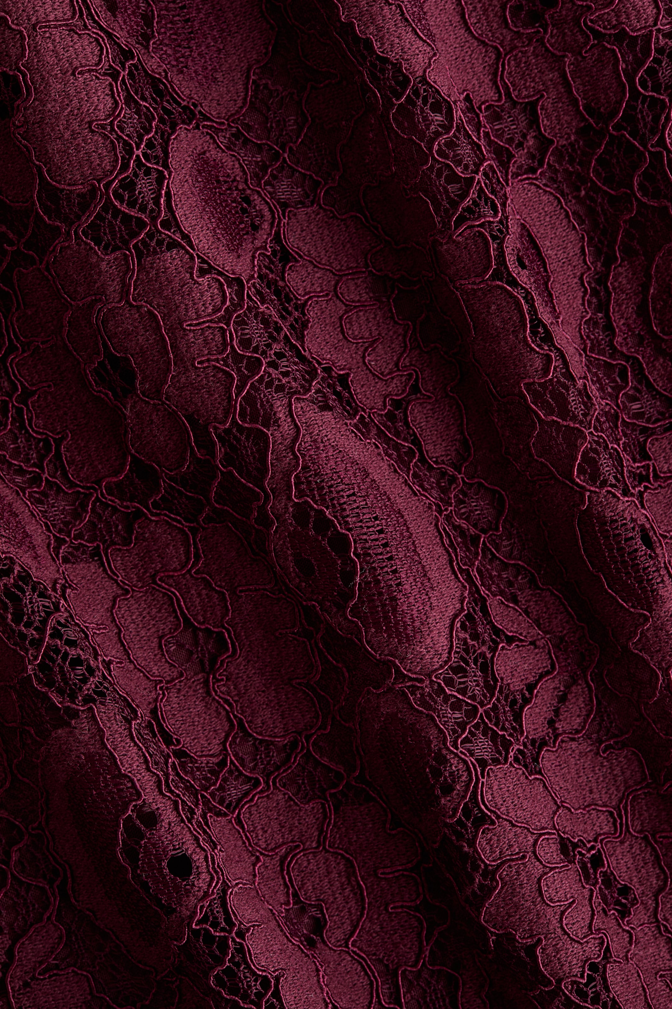 Ampliar la imagen: La tela de encaje exhibe un color marrón oscuro profundo, con patrones florales intrincados que se superponen sobre una base sólida. La tela está drapeada, creando pliegues suaves que enfatizan su textura y diseño.