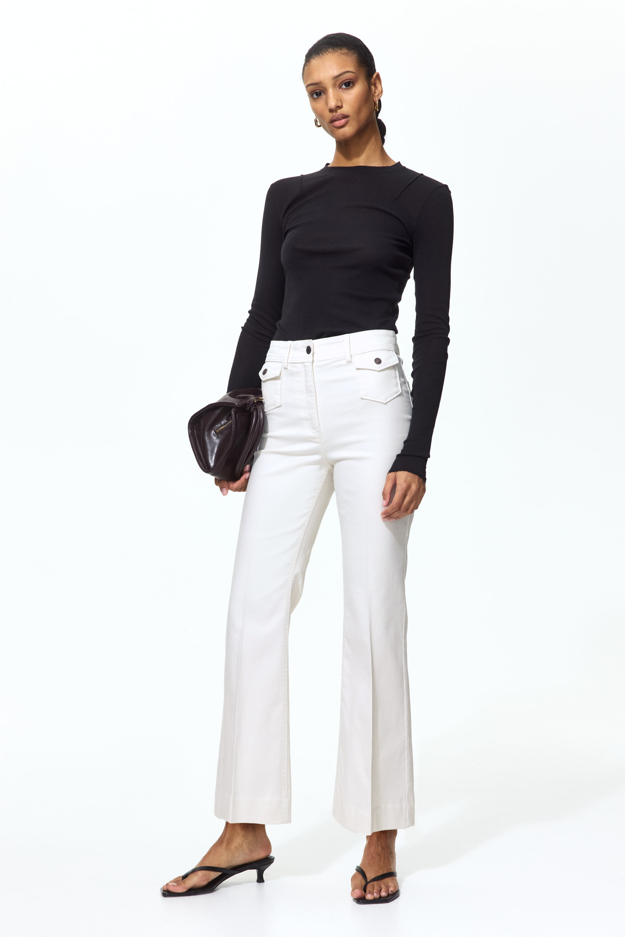 View larger image: The model is wearing fitted white twill trousers, characterized by a high waist with a button closure, patch front pockets with buttoned flaps, and flared legs with front creases. Paired with a black long-sleeved top, these trousers offer a chic and sophisticated look.