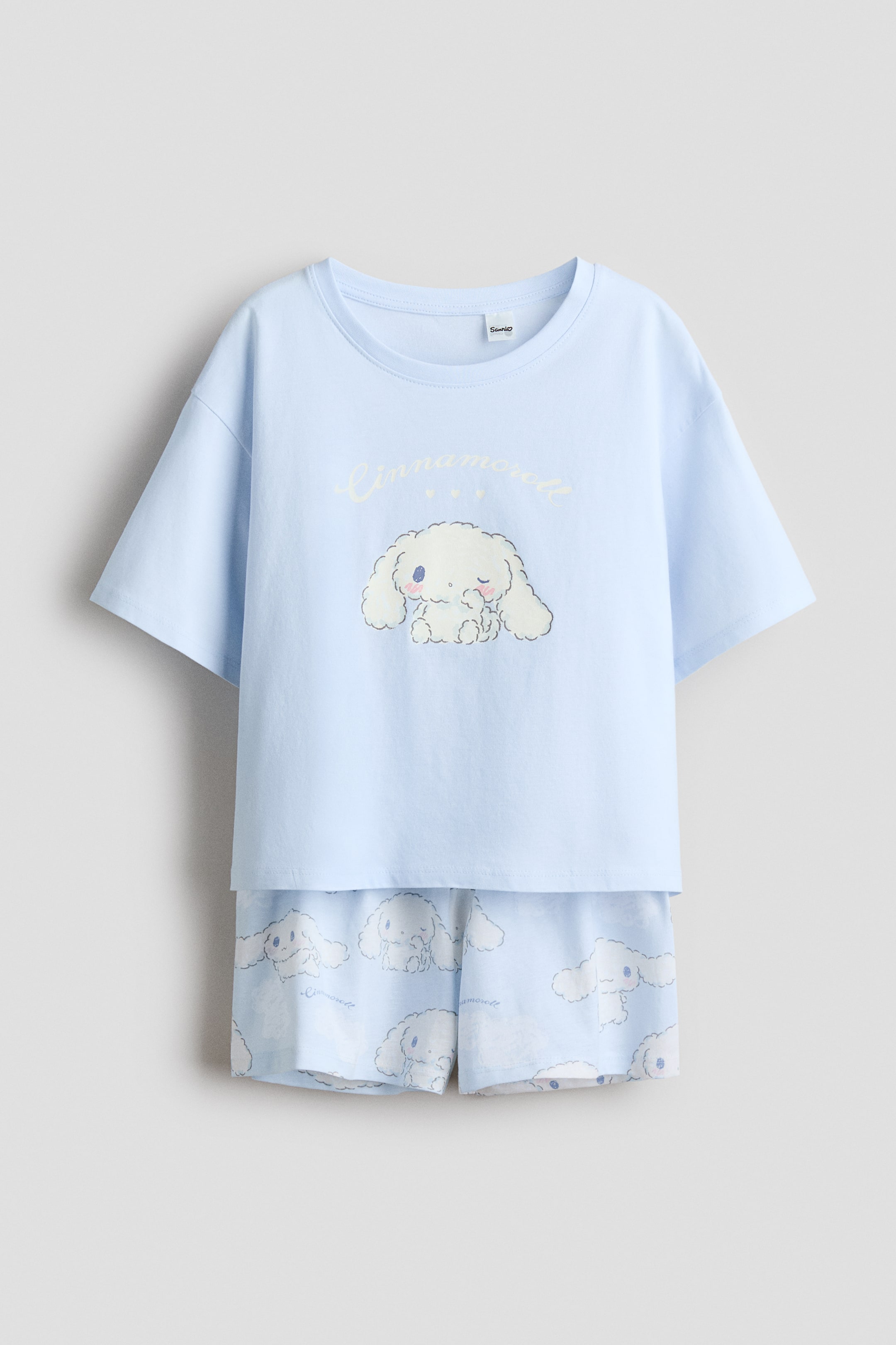 View larger image: A front view of light blue cotton jersey pyjamas, featuring a short-sleeved t-shirt and matching shorts. The t-shirt boasts a ribbed trim around the neckline, dropped shoulders, and a large white Cinnamoroll character print with 'Cinnamoroll' text on the front. The shorts display an all-over print of smaller Cinnamoroll characters, clouds, and text, designed with an elasticated waist.