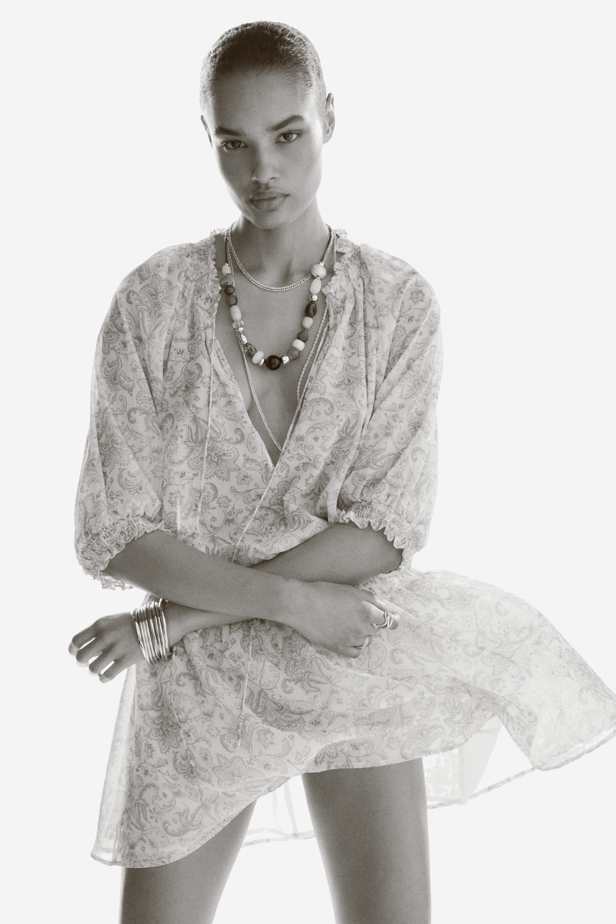 View larger image: The model is showcasing a short, oversized tunic dress crafted from a patterned woven fabric. It features a smocked round neckline with a V-shaped opening and delicate spaghetti ties. The dress also boasts short, voluminous raglan sleeves with smocking at the cuffs and a smocked waistband that transitions into a gently flowing flared skirt. The model has her arms crossed, accentuating the dress with an array of necklaces, bracelets, and rings.