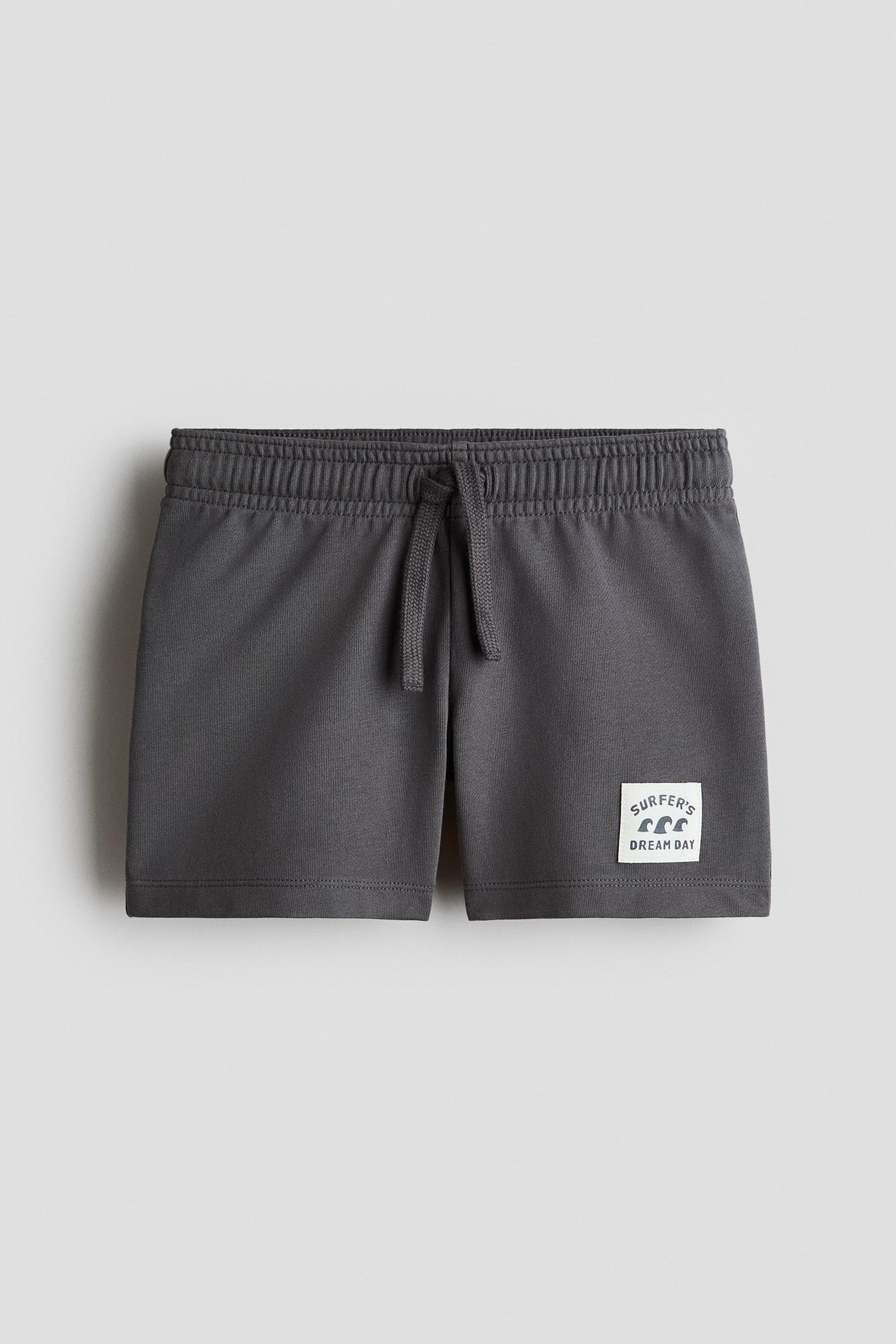View larger image: Loose-fit shorts in dark grey lightweight cotton sweatshirt fabric are showcased from the front, featuring an elasticated, drawstring waist with the drawstring tied. A white patch with 'SURFER'S DREAM DAY' text and wave symbols appears on the lower right leg.