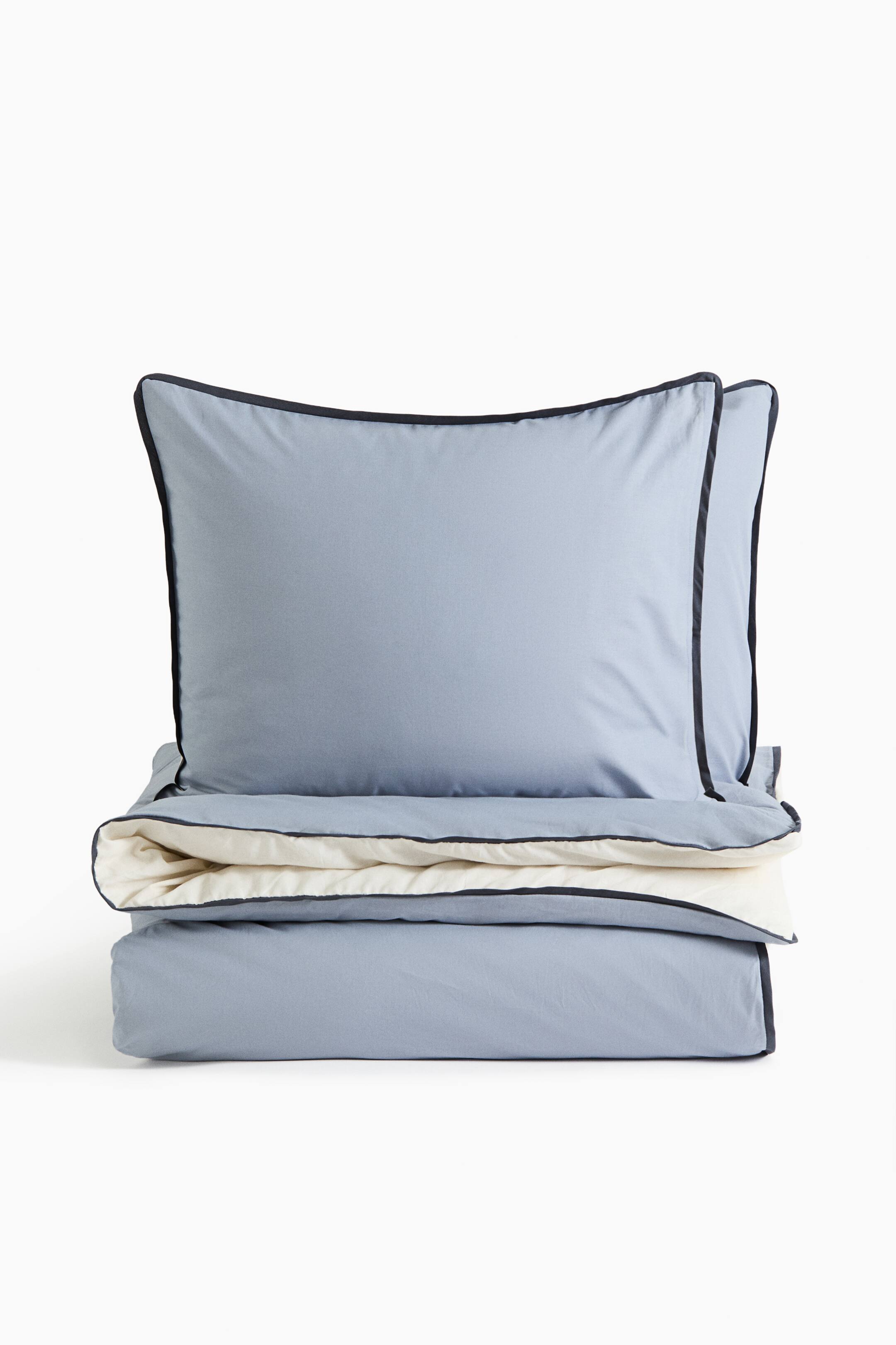 Agrandir l'image: Un ensemble de literie présente une housse de couette bleu clair et deux cache-oreillers assortis, maintenus debout et tournés vers l’avant. La housse de couette pliée laisse apparaître un envers contrasté de couleur crème, et les deux pièces sont bordées d’un fin liseré passepoilé bleu marine foncé le long des bords.