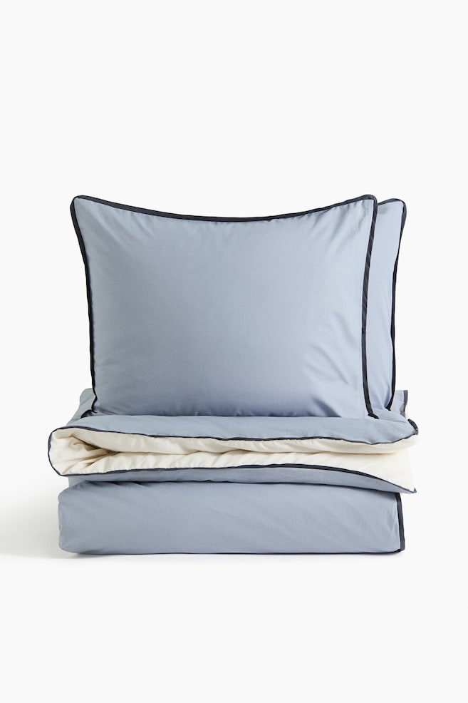 A bedding set is displaying a light blue duvet cover and two matching pillow shams propped upright facing front. The folded duvet cover is showing a contrasting cream-coloured underside, with both pieces featuring a thin, dark navy piped border along the edges. - 1