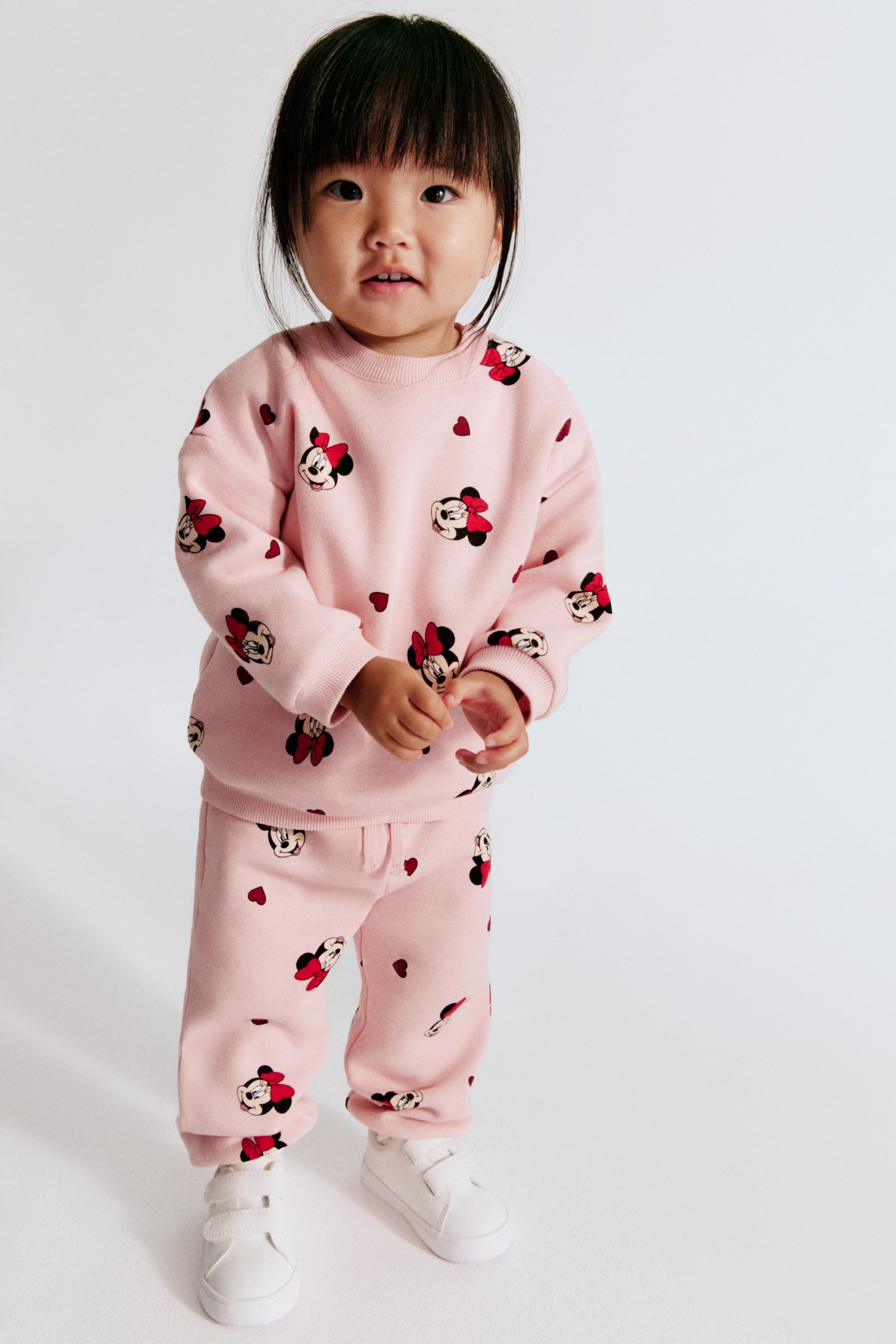 View larger image: A 2-piece print sweat fabric set in light pink is shown from the front, featuring an all-over pattern of Minnie Mouse faces with red bows and small red hearts. The set includes a long-sleeve sweatshirt with a ribbed crew neck and cuffs, paired with matching sweatpants with gathered ankles.