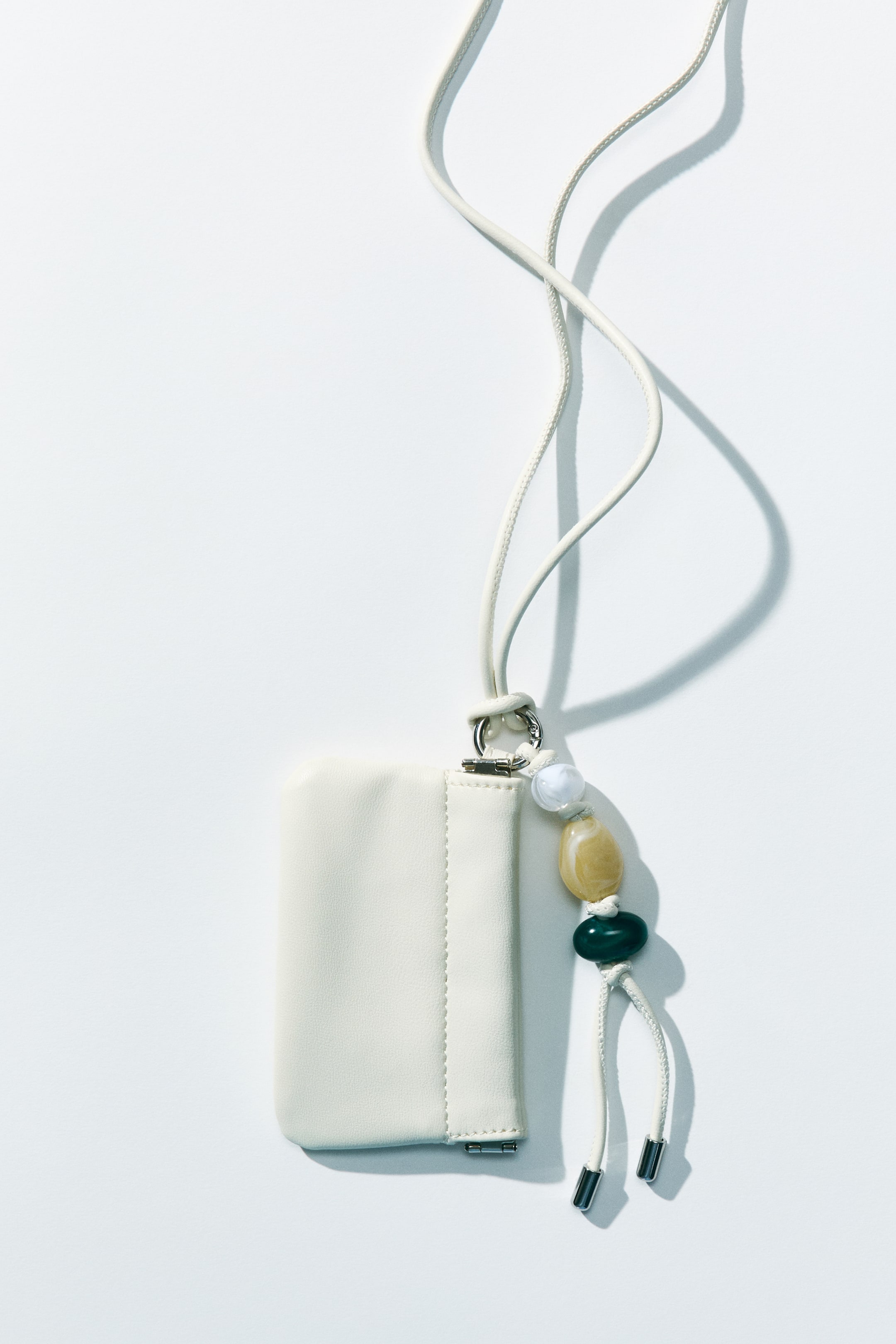 Zobacz większy obraz: Kremowa mini torebka crossbody z powlekanej tkaniny leży płasko, widziana od przodu, z wąskim paskiem na ramię unoszącym się z srebrnego pierścienia. Wzdłuż górnej krawędzi widoczne jest ukryte metalowe zapięcie ramy, a do tego samego pierścienia za pomocą sprężynowego zapięcia przypięty jest ozdobny brelok z białymi, żółtymi i ciemnozielonymi koralikami oraz sznurkami zakończonymi srebrnymi frędzlami.