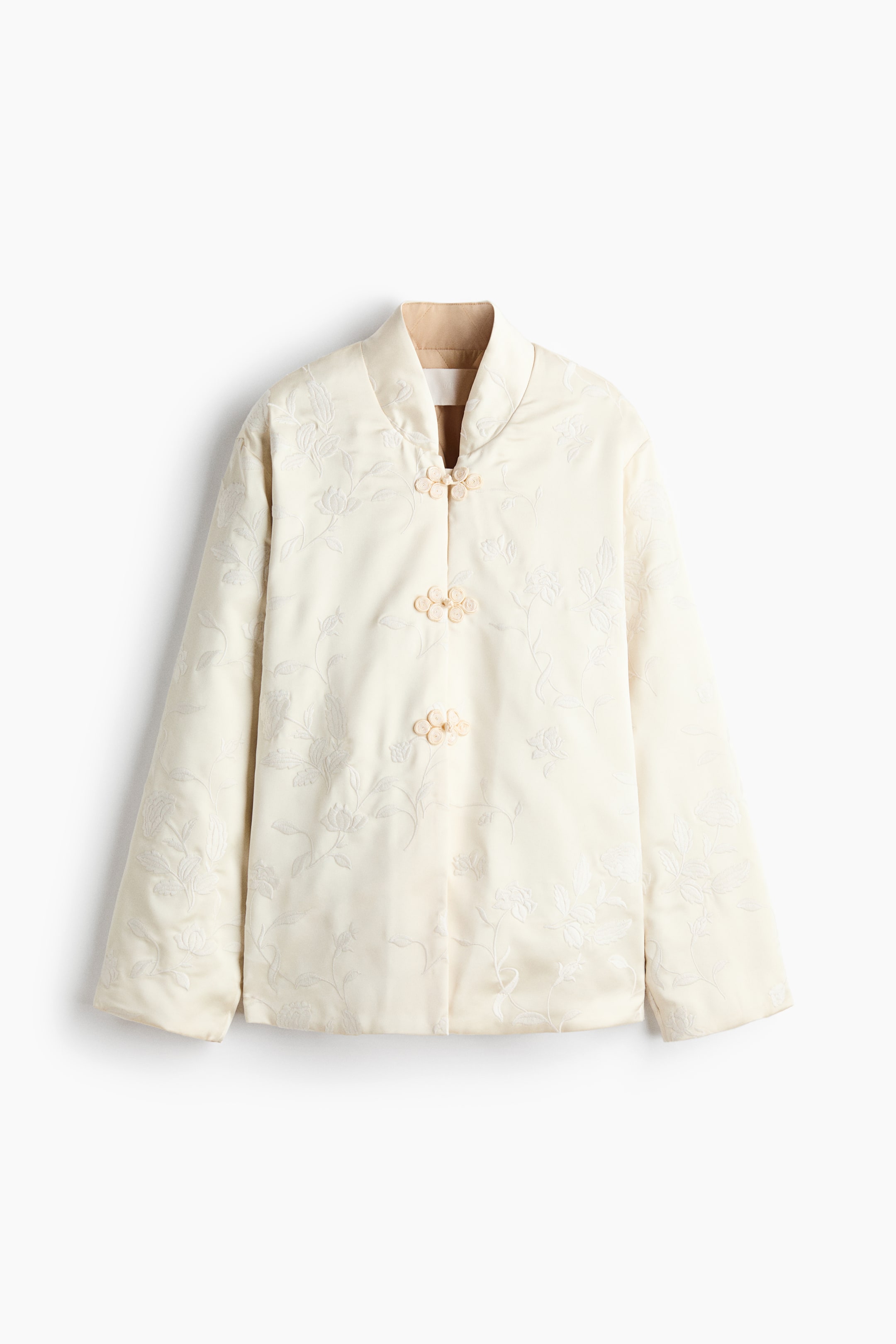View larger image: A cream-colored padded jacket is shown from the front, featuring a subtle floral jacquard pattern and a stand-up collar with contrasting tan lining. The jacket has three decorative fabric knot closures down the center front and long, wide sleeves.