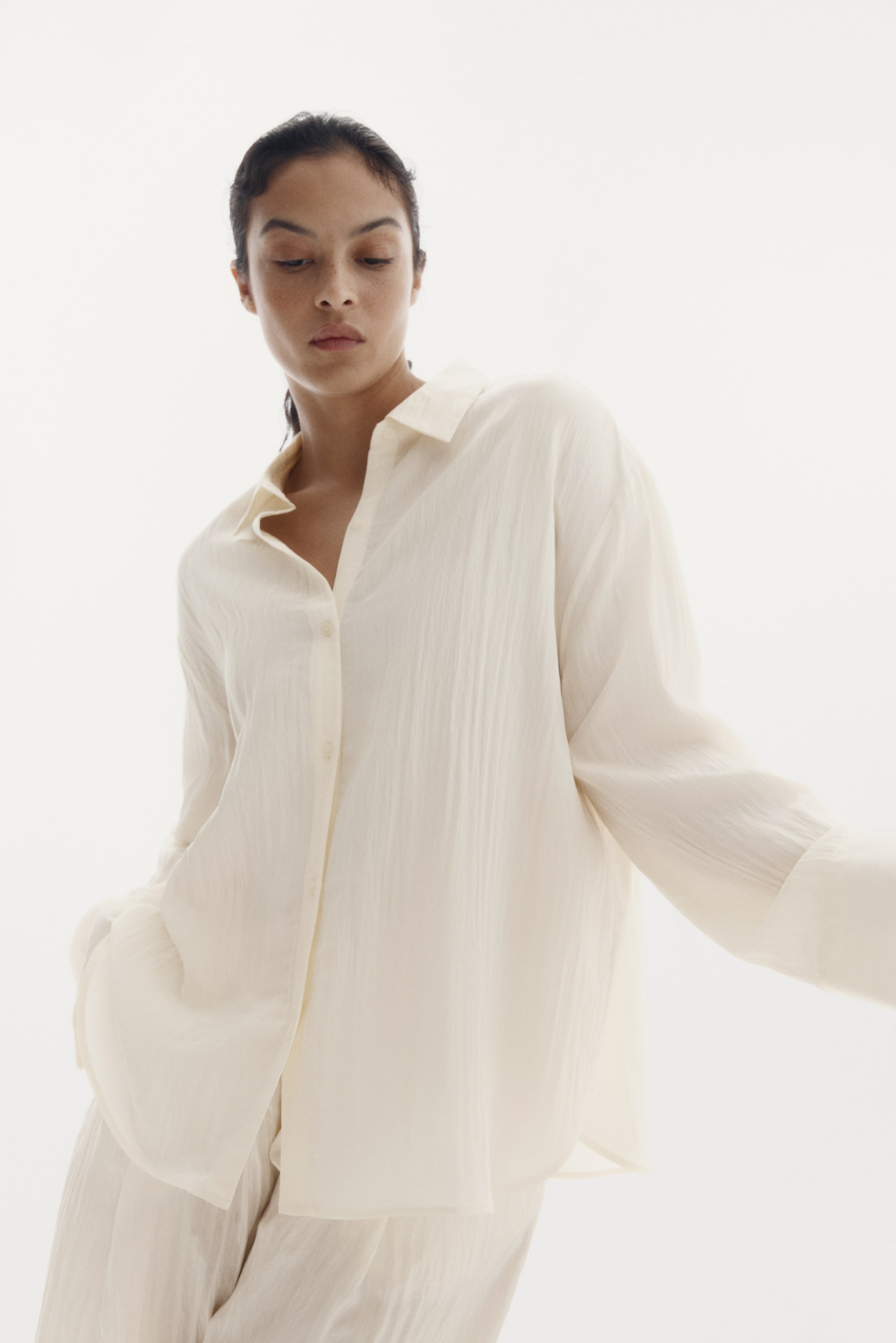 Zobacz większy obraz: Kremowa koszula z zapięciem na guziki charakteryzuje się pomarszczoną teksturą, otwartym kołnierzem oraz długimi, luźnymi rękawami. Jest noszona w swobodnym kroju, skierowana do przodu i lekko pochylona w dół, zestawiona z pasującymi kremowymi spodniami.