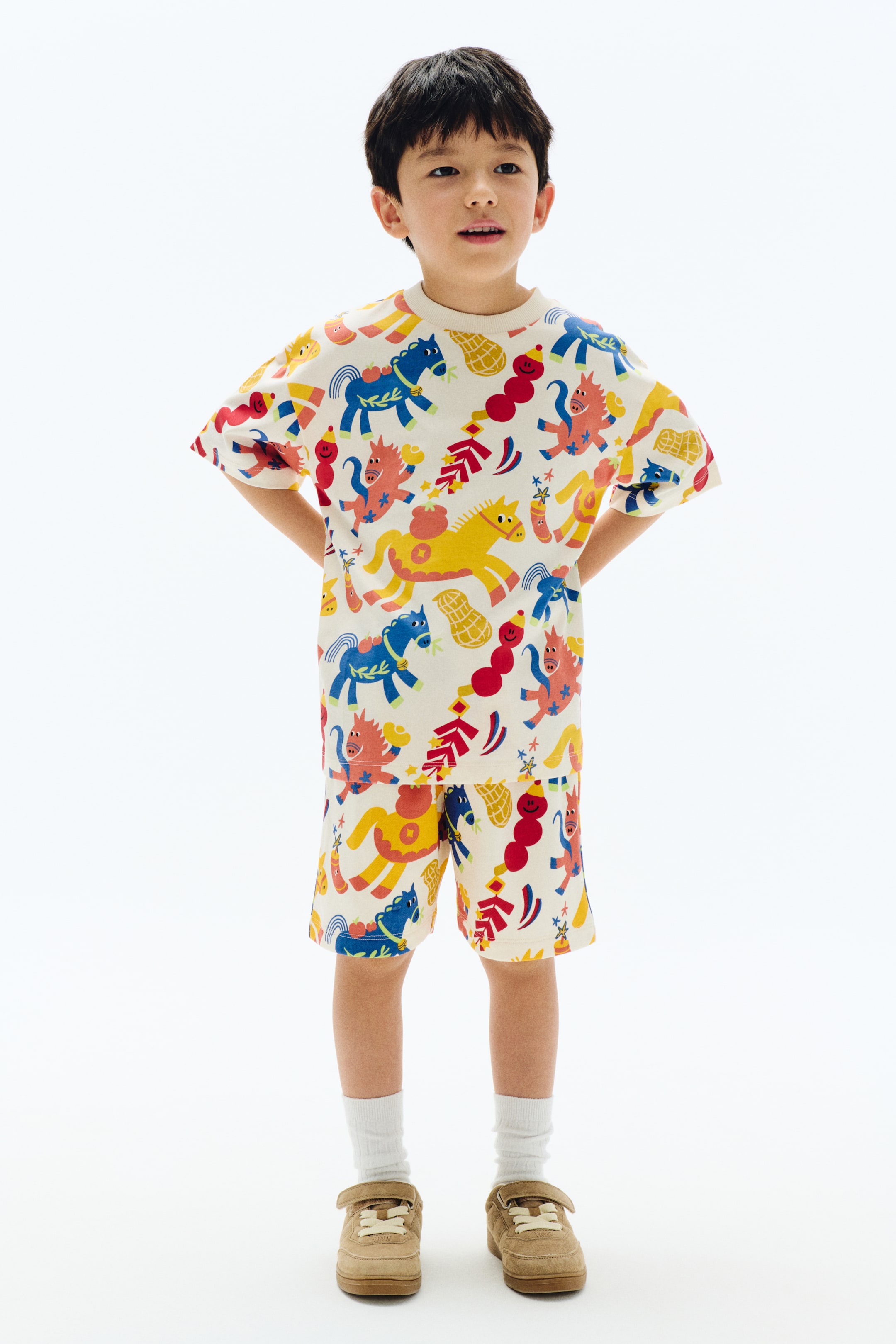 View larger image: A two-piece cream-colored outfit, consisting of a t-shirt and matching shorts, features an all-over colorful pattern of stylized horses, peanuts, and abstract shapes. The t-shirt has a crew neck and short sleeves, while the shorts fall above the knee, showing a full front view of the set.