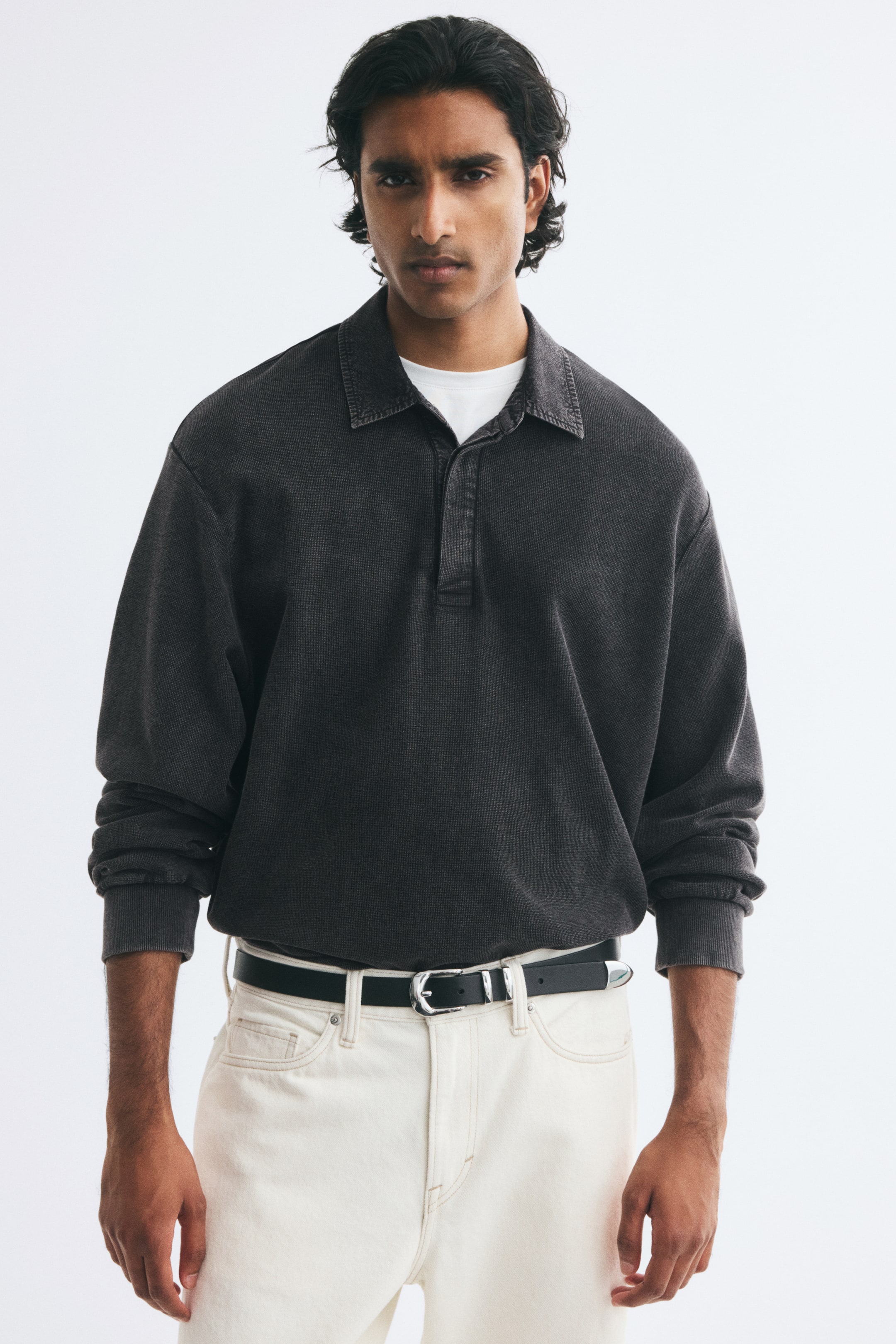 View larger image: A dark grey long-sleeved polo shirt with a washed finish is being worn by the model, layered over a white crew-neck T-shirt and tucked into off-white trousers with a black belt. The shirt features a relaxed fit with dropped shoulders, ribbed cuffs, a collar, and a concealed button placket.