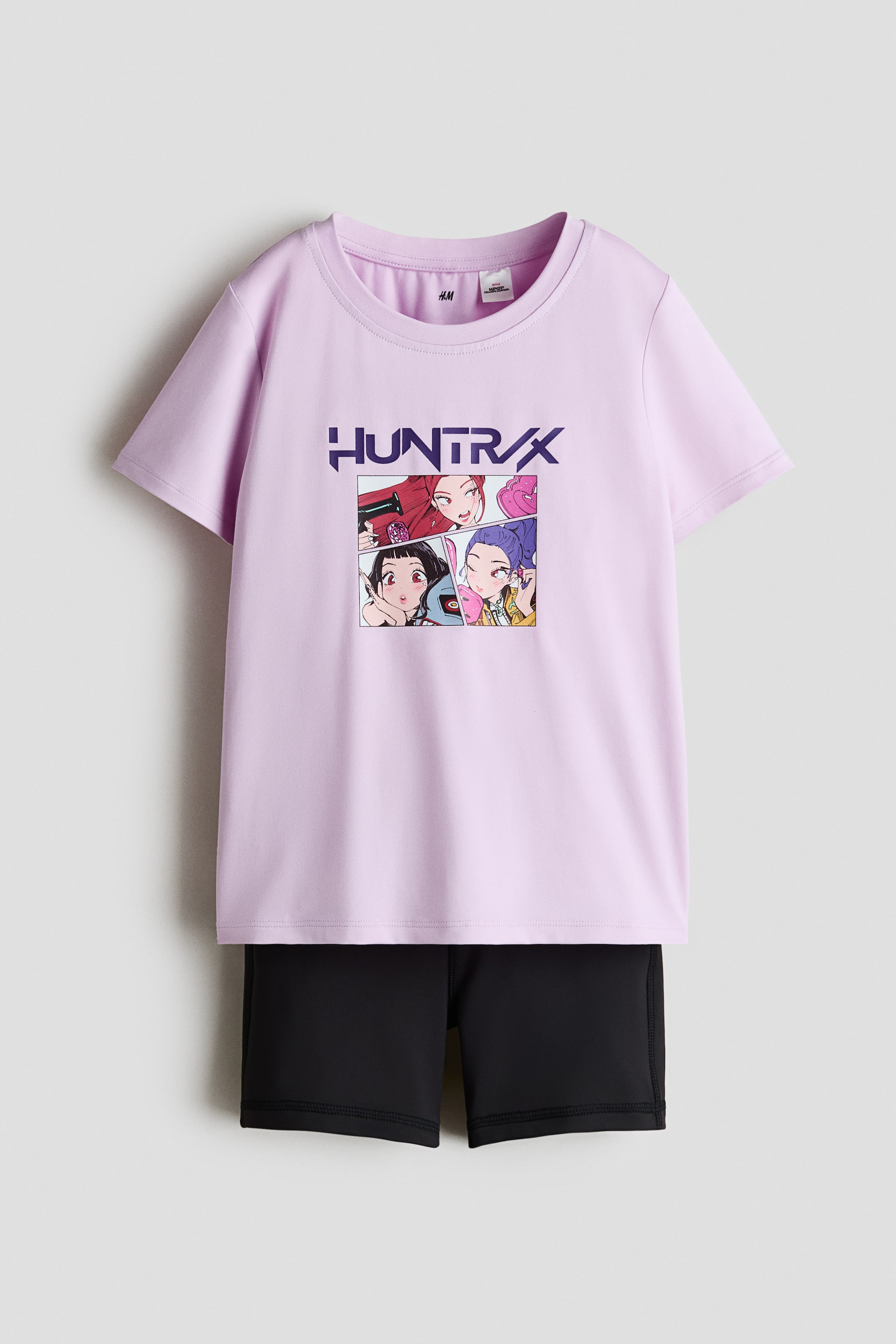 View larger image: A light purple short-sleeved t-shirt with a crew neck showcases a central graphic print on the front, displaying the text "HUNTR/X" above a four-panel comic-style illustration of female animated characters. The t-shirt is paired with black shorts worn underneath.